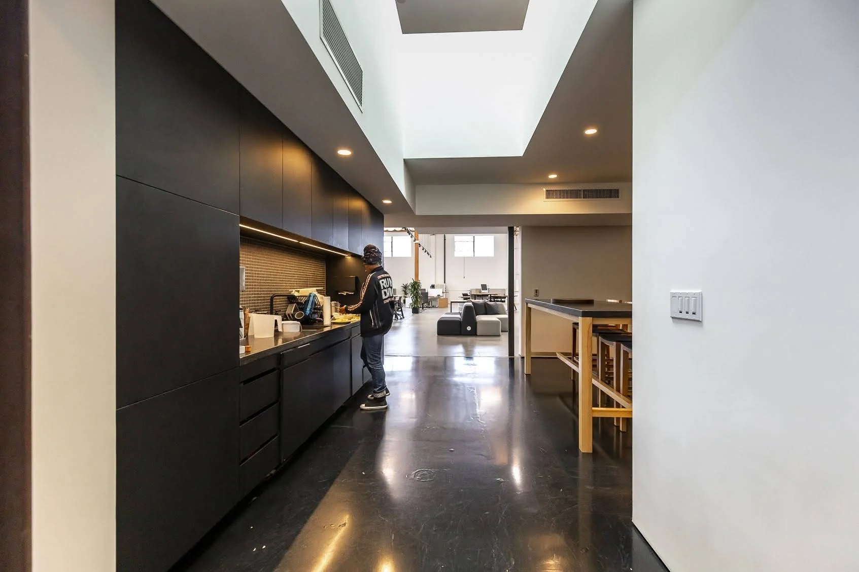 Interior of a modern kitchen with black cabinets, a person preparing food, and a living room with couches and a large window in the background.