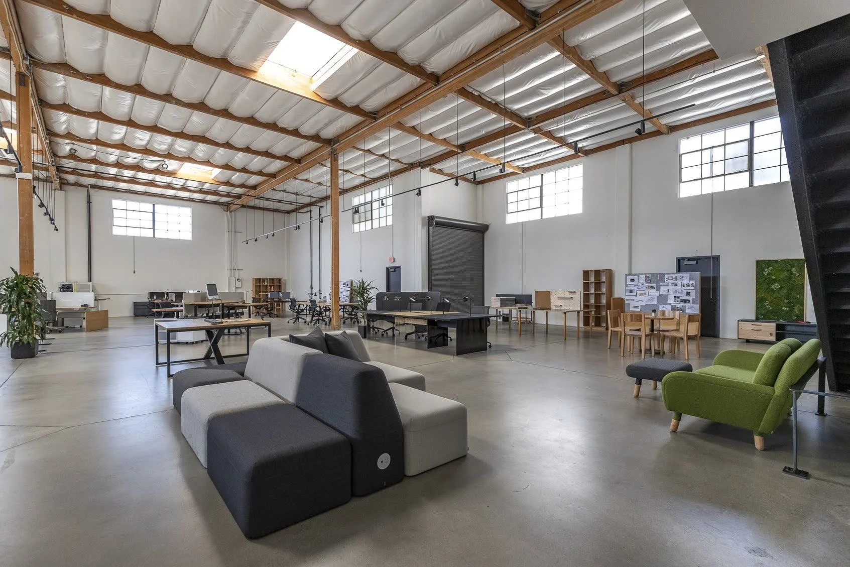 An spacious industrial-style office with high ceilings, large windows, and a variety of furniture including desks, chairs, tables, and sofas.