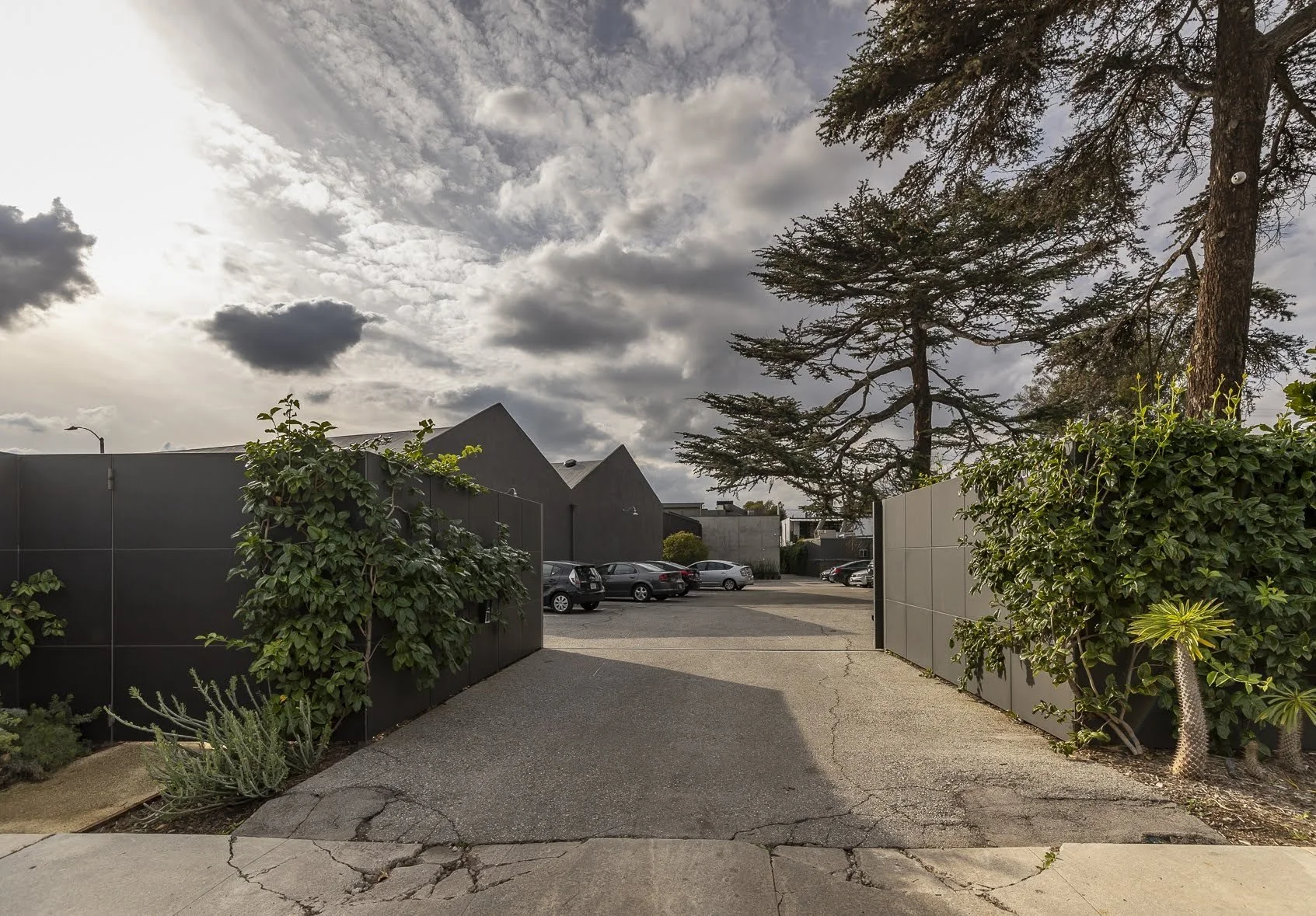 View of a parking lot surrounded by black and beige fences, with a few cars parked and tall trees, under a cloudy sky.