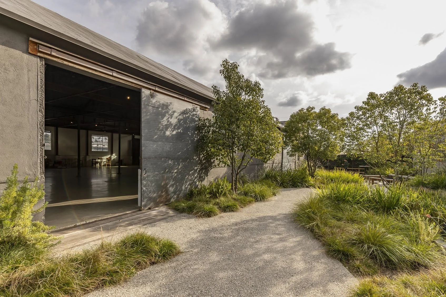 Outdoor courtyard with a gravel pathway, trees, and lush green plants, adjacent to a modern building with an open gray metal garage door