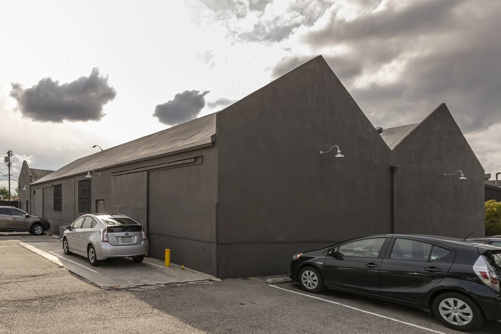 A dark gray building with a steep roof, outdoor wall-mounted lights, and parked cars in front, with a cloudy sky overhead.