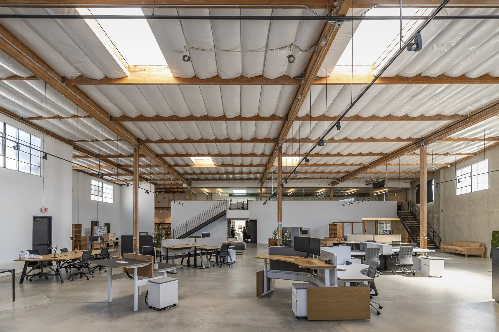 Open modern industrial-style office space with high ceilings, large windows, work desks, chairs, and a staircase leading to an upper level.