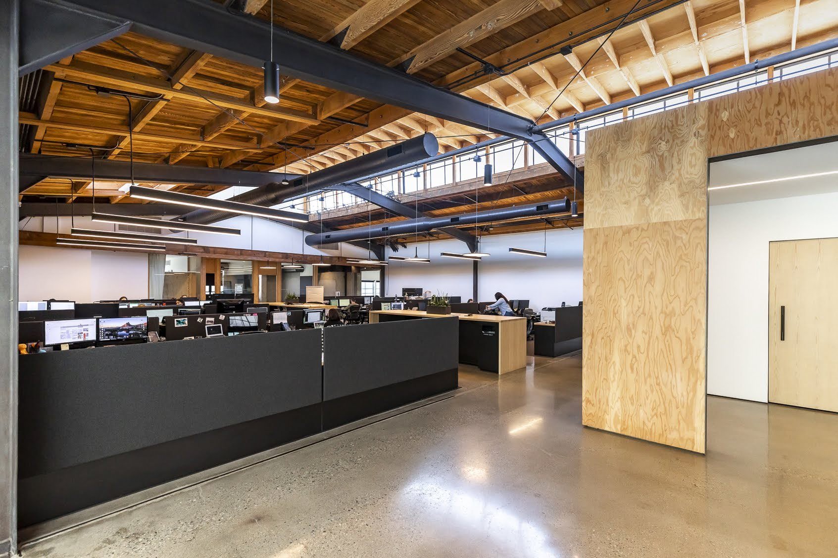 Open-plan modern office with exposed wooden ceiling beams, black ductwork, and multiple workstations with computers and monitors.