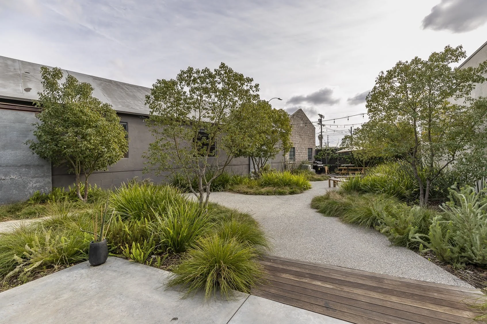 A landscaped urban backyard with a gravel walking path, surrounded by trees, grass, and various plants, with patio seating and string lights in the background.