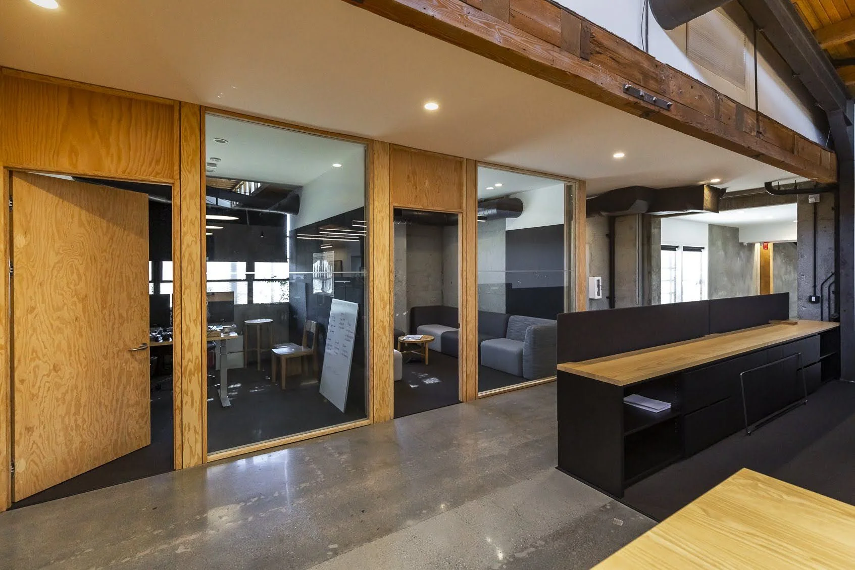 Office interior with glass partitioned rooms, wooden and black furniture, and concrete accents.