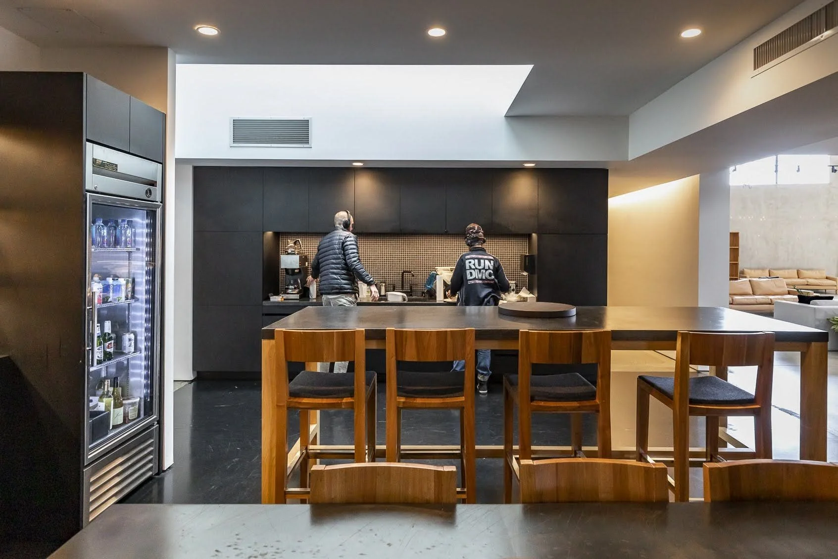 Two people working behind a coffee bar in a modern cafe, with a wooden counter and chairs in the foreground.