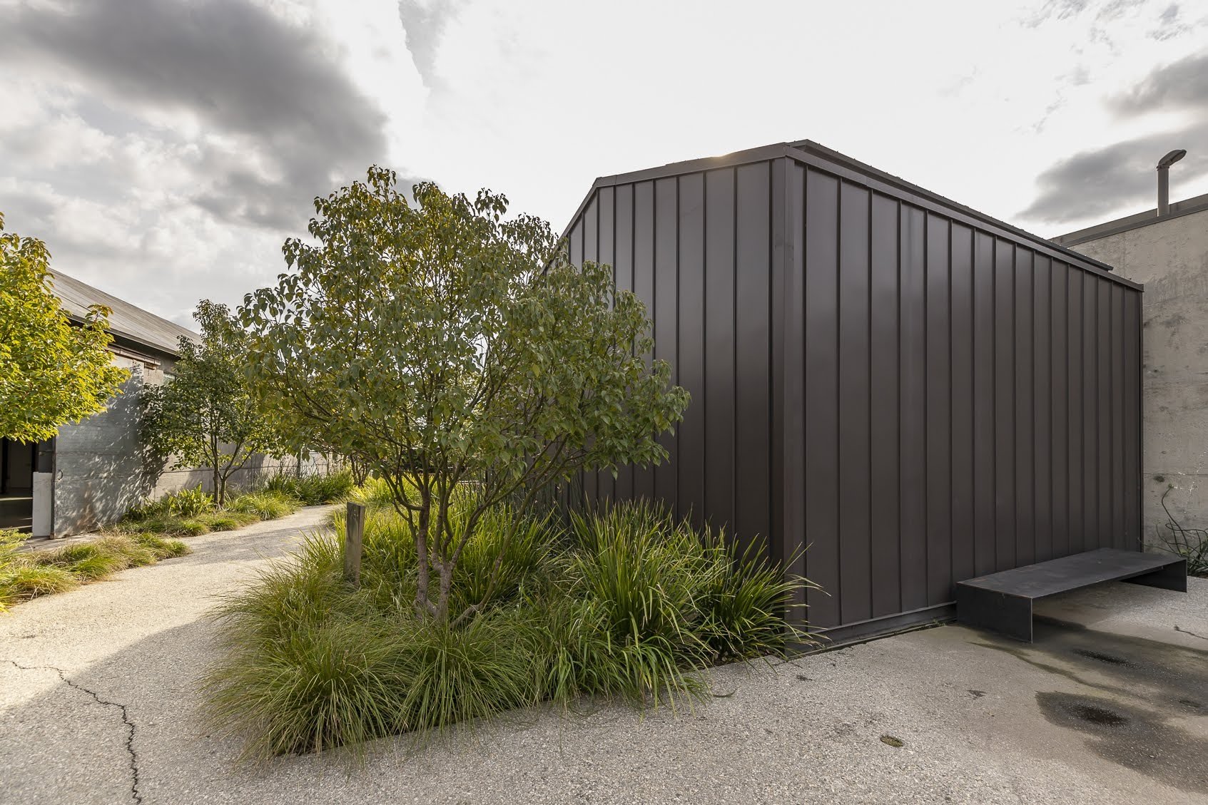 A modern building with dark metal siding, surrounded by green bushes and trees, with a cracked pavement and a cloudy sky.