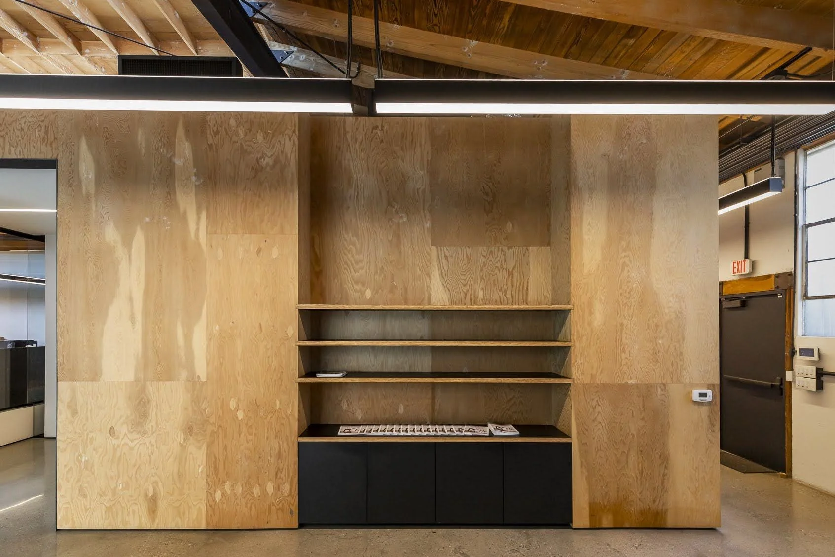 Interior of a modern building with wood-paneled walls, open shelving, and ceiling beams, with partial view of a hallway and door on the right.