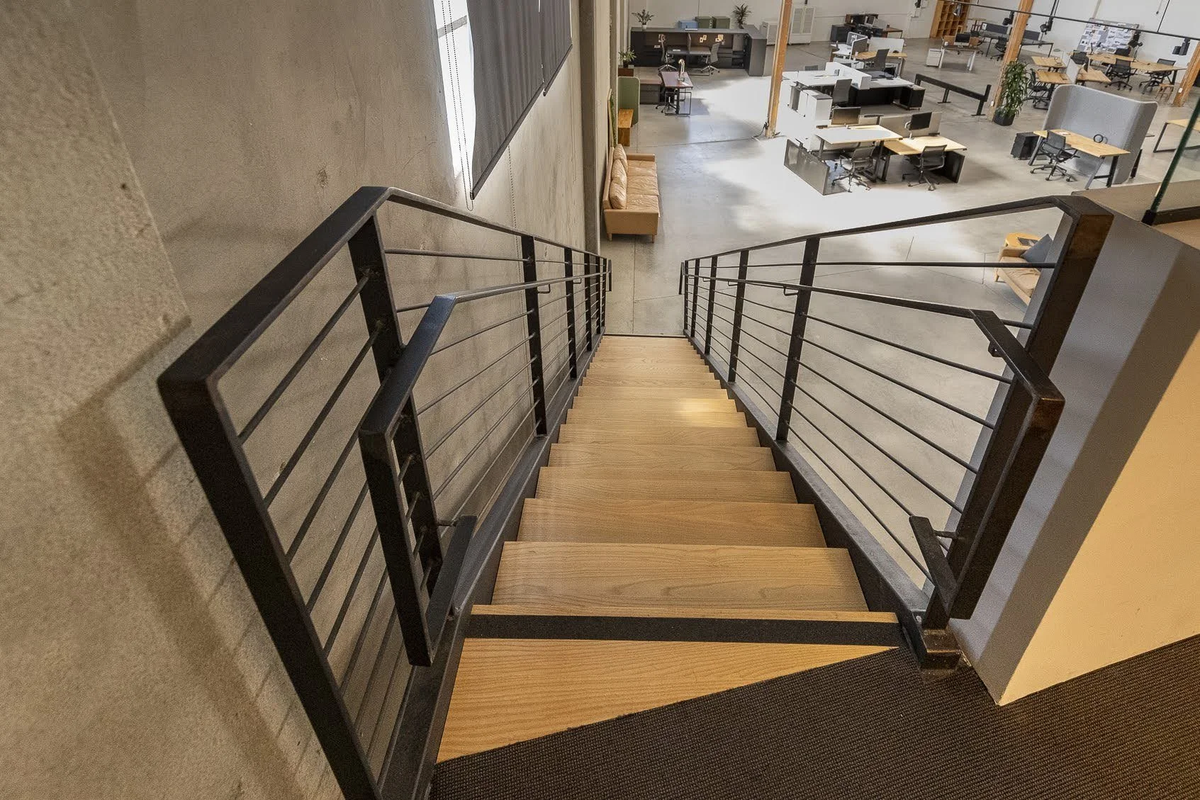 A staircase with wooden steps and black metal handrails leading down to an open office space with desks and chairs.