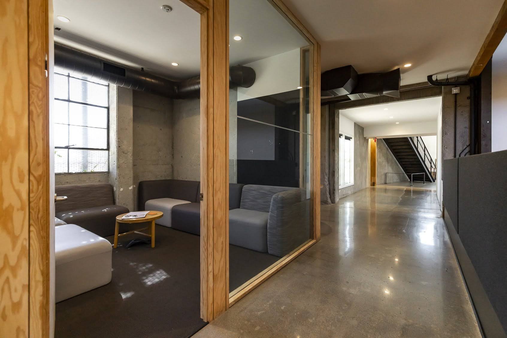 View of a modern, minimalist lobby area with concrete walls, wooden accents, and a mix of seating arrangements visible through glass partitions, leading to a staircase in the background.