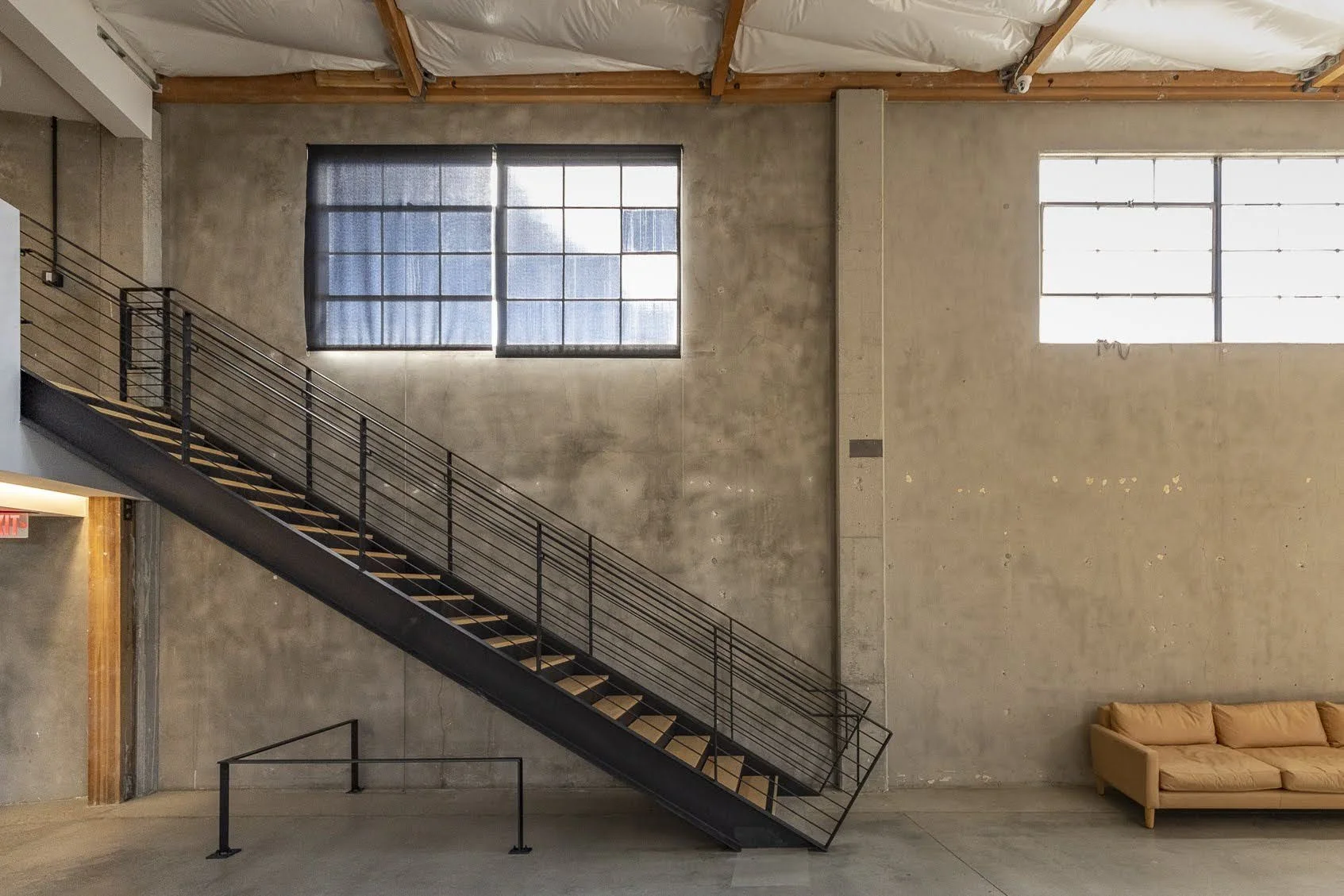 Interior view of an industrial-style space with a black metal staircase, large windows with black framing, exposed concrete walls, and a tan sofa to the right.