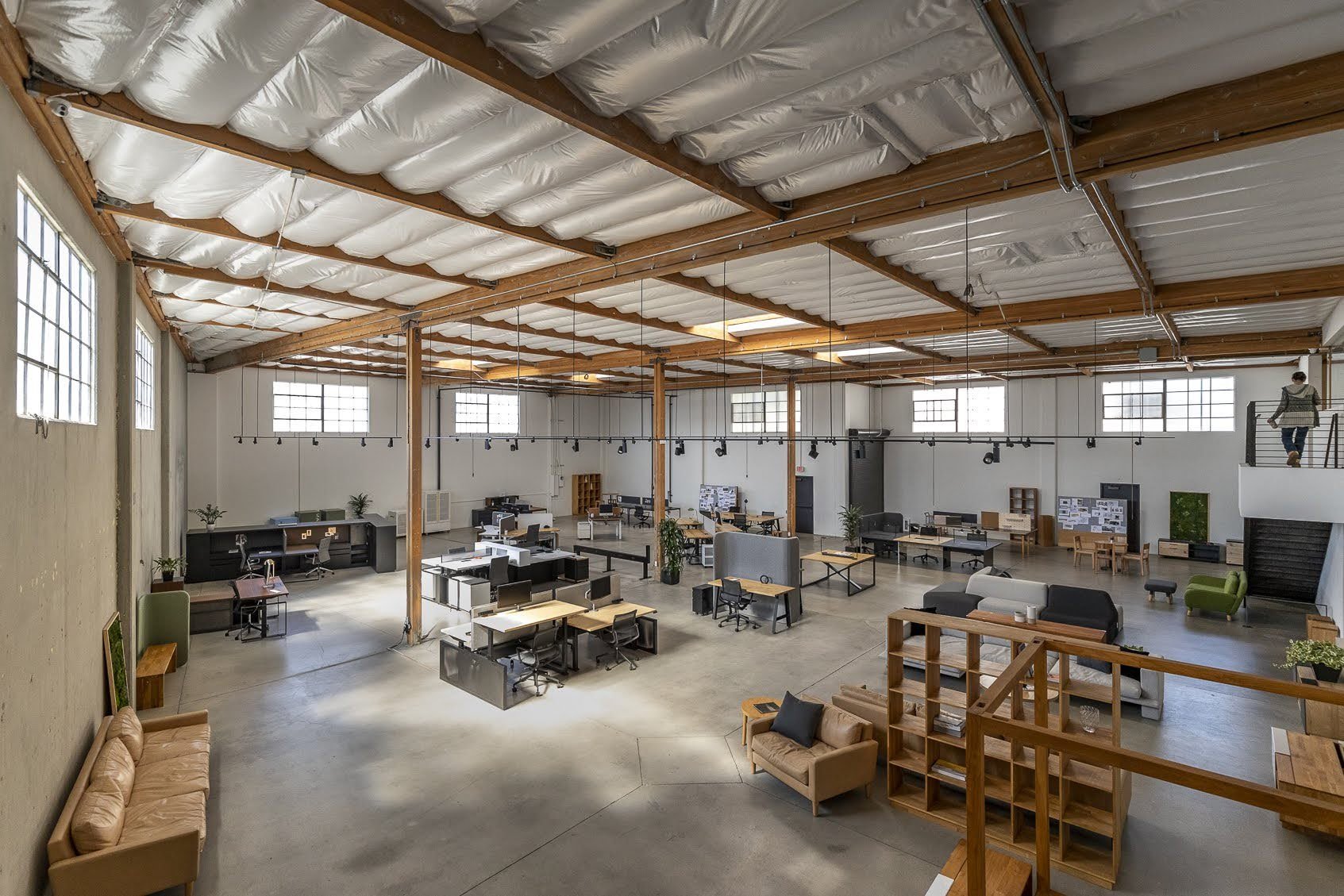 A spacious modern office with high ceilings, large windows, multiple workstations, lounge seating, and a person on the upper level staircase.