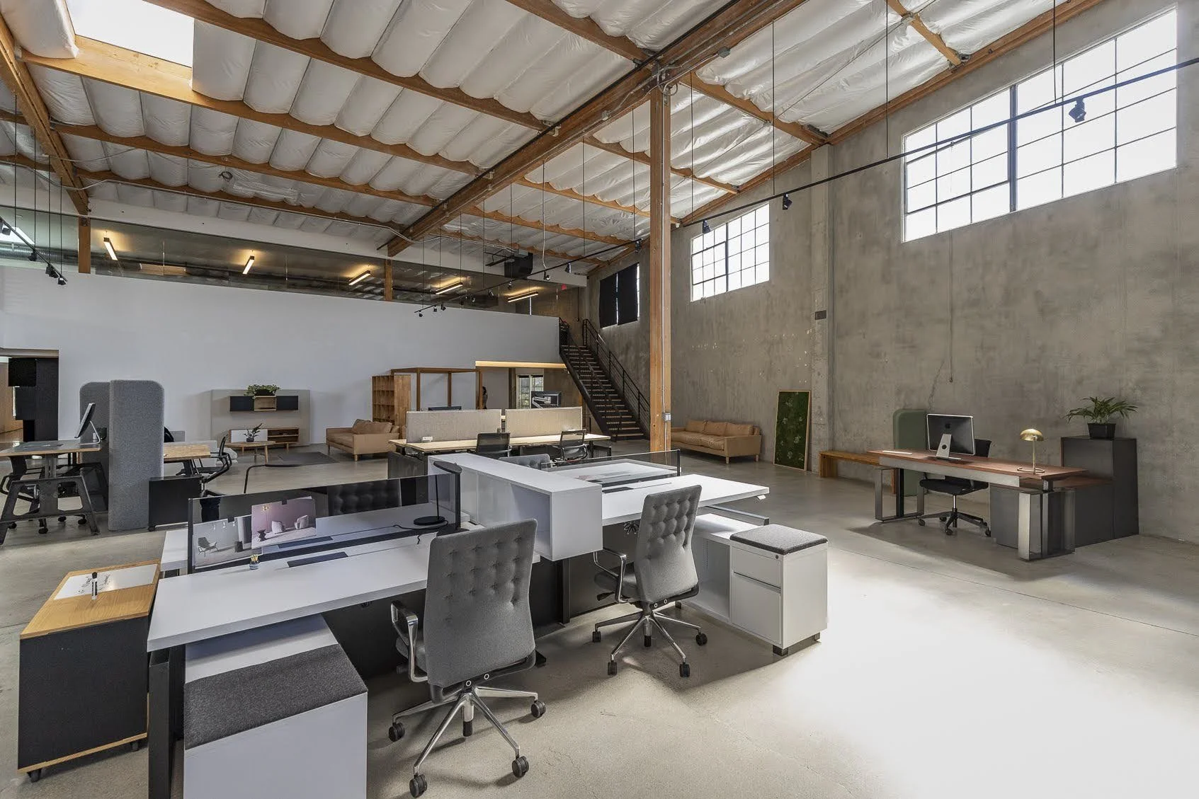 Open-concept coworking space with multiple desks, chairs, and lounge areas, high ceilings with exposed beams, large windows, and a concrete wall.
