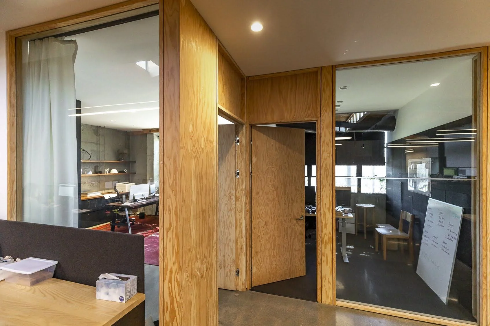 Interior office space with wooden framed glass walls, showing a conference room with chairs, a whiteboard with writing, and desks with computers.
