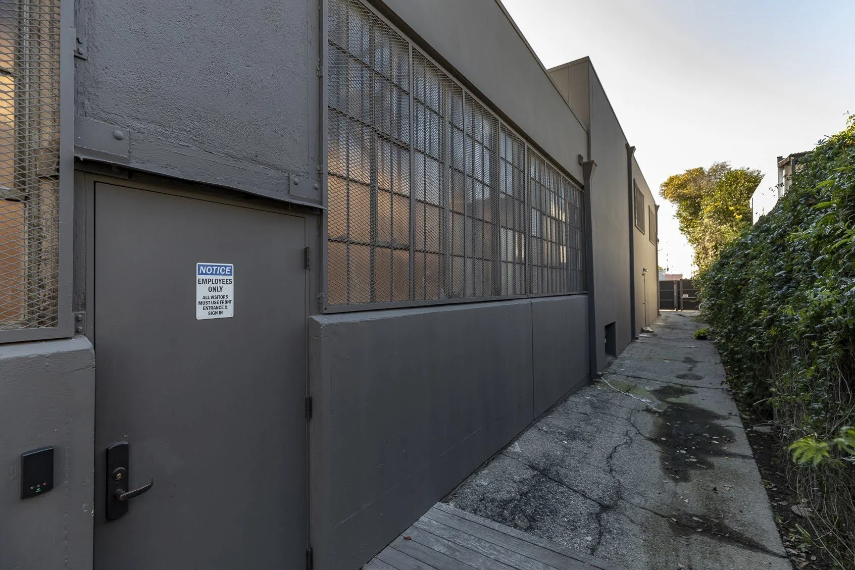 Back alley with a gray metal door that has a sign indicating employees only. The alley is cracked with patches and is bordered by a gray building on one side and green shrubbery on the other. Trees are visible in the distance under a clear sky.