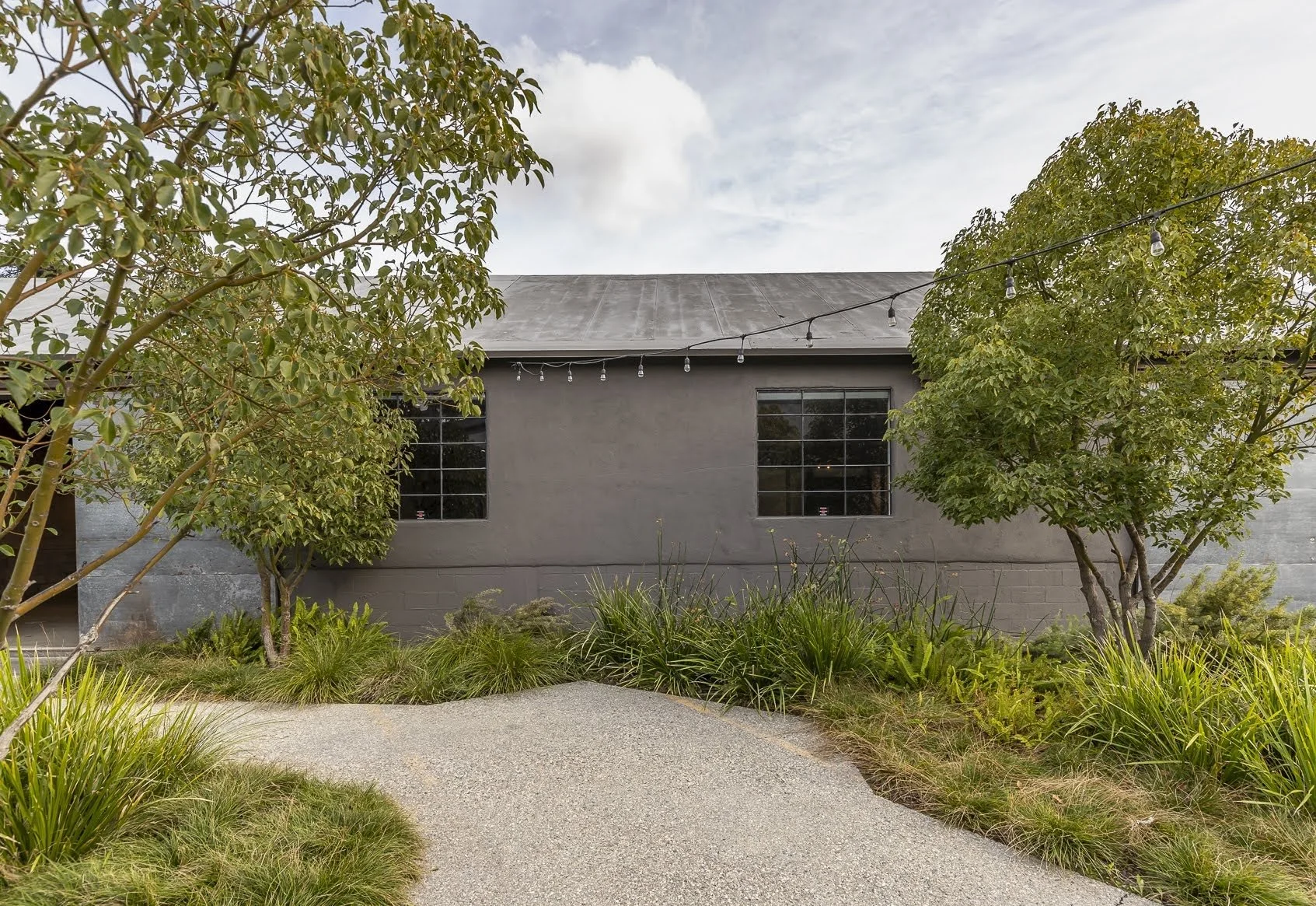 Exterior view of a modern building with gray walls, large black-framed windows, surrounded by greenery, trees, and a gravel pathway.