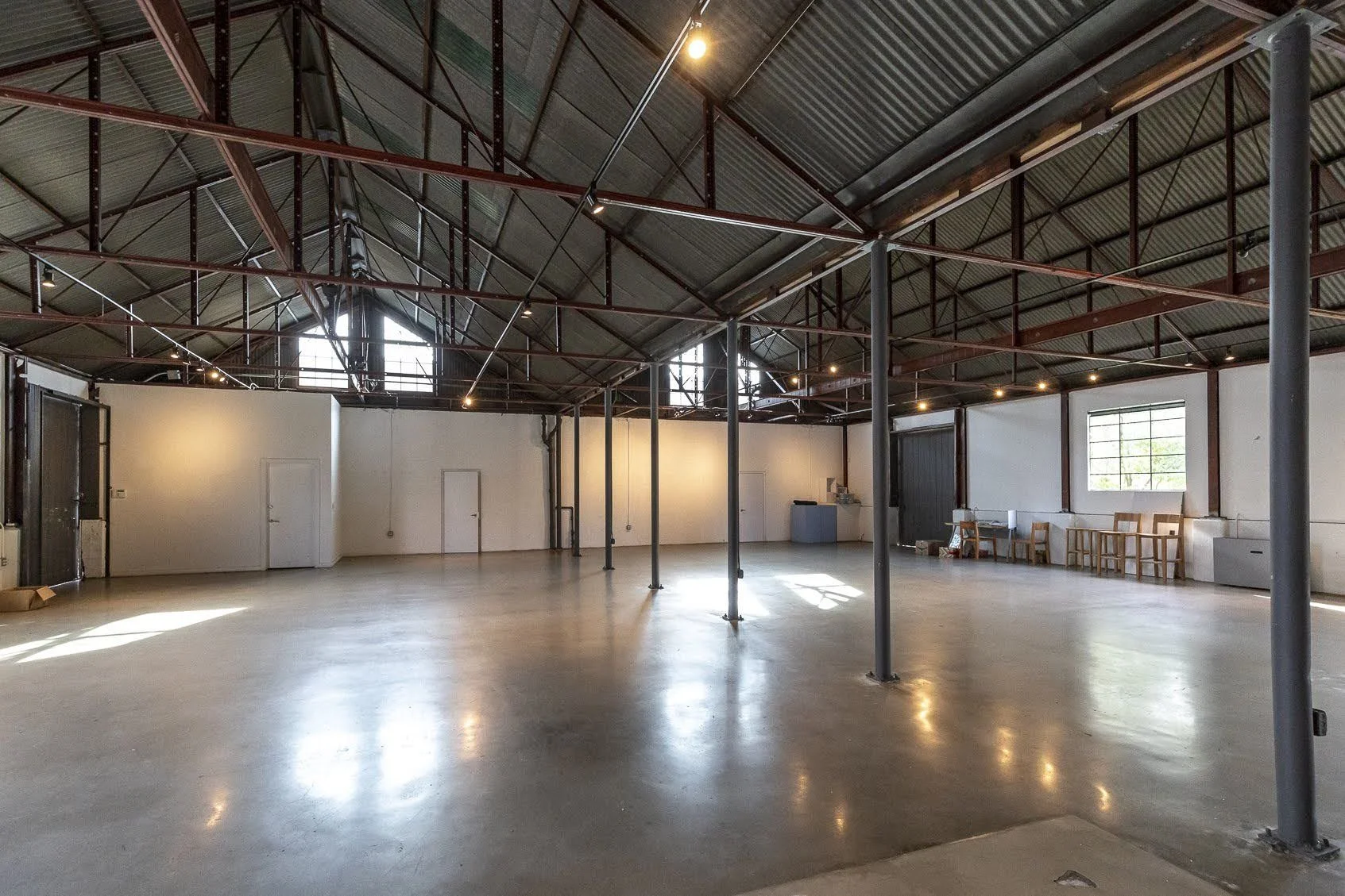 Empty industrial-style warehouse with high metal roof, open floor space, and a few chairs near the window.