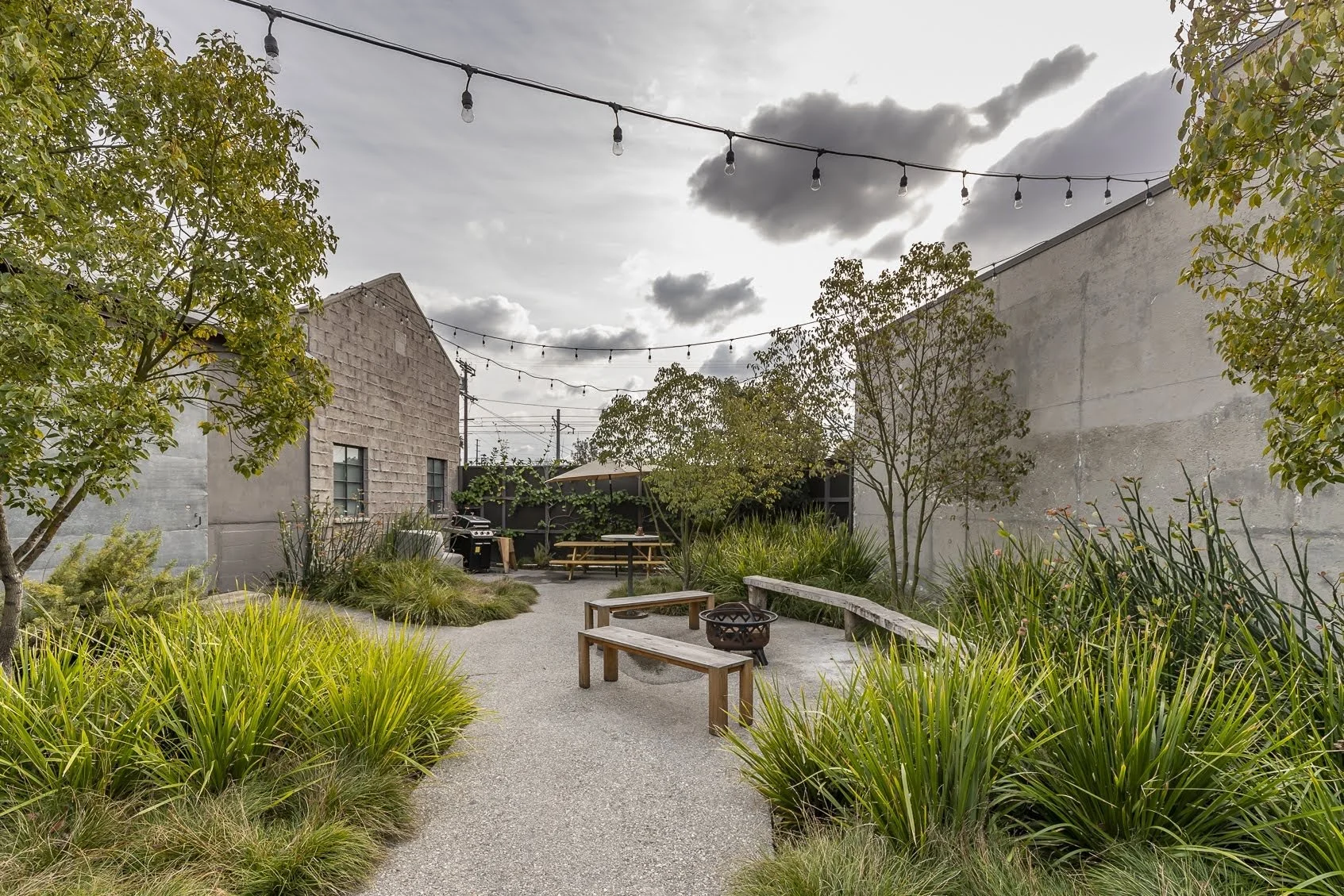 A cozy outdoor patio area with a gravel pathway, surrounded by green plants and trees. Wooden benches and a fire pit are visible. String lights are hung overhead under a partly cloudy sky.