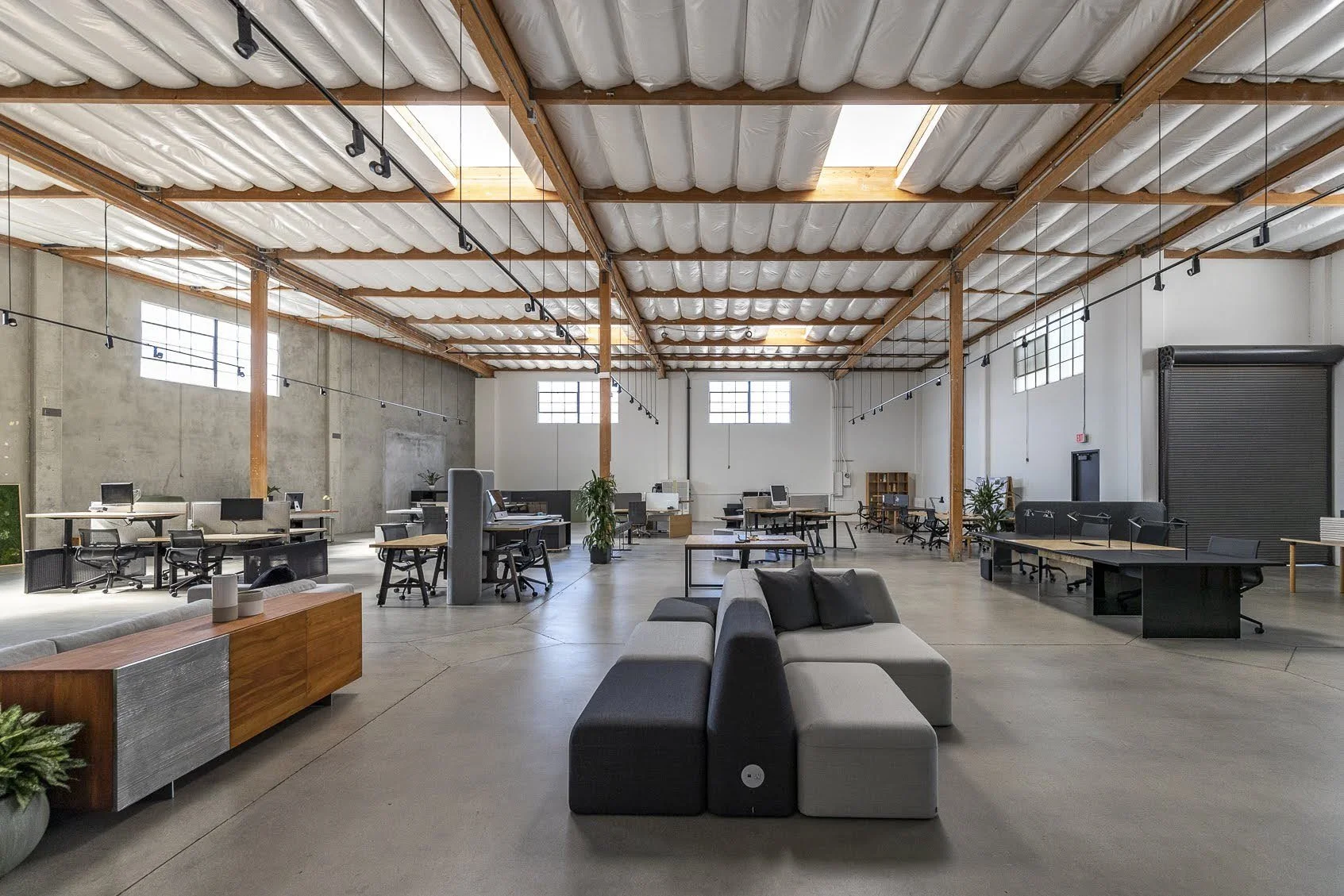 A spacious, modern open-concept office with high ceilings, exposed wooden beams, large industrial windows, and various workstations including desks with computers, a seating area with a sectional sofa, and potted plants.