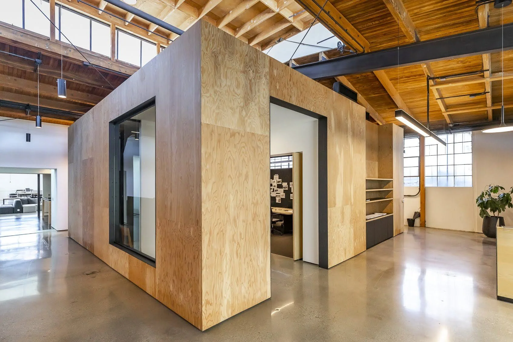 Interior of a modern office space with wood-paneled walls, large windows, and exposed ceiling beams. There is a small enclosed room with glass and wood walls, open shelving, and a plant in a black pot.