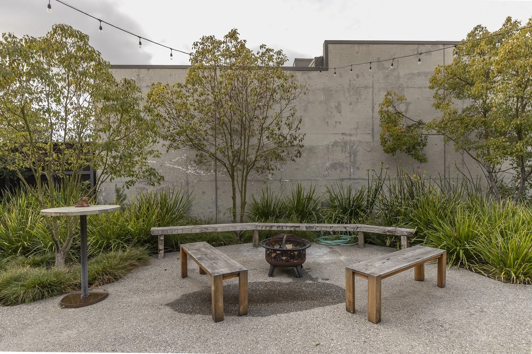 A small outdoor patio with a concrete floor, a wooden bench, two wooden stools, a fire pit, a metal table with a small object on it, green plants, trees, a gray wall, and string lights overhead.