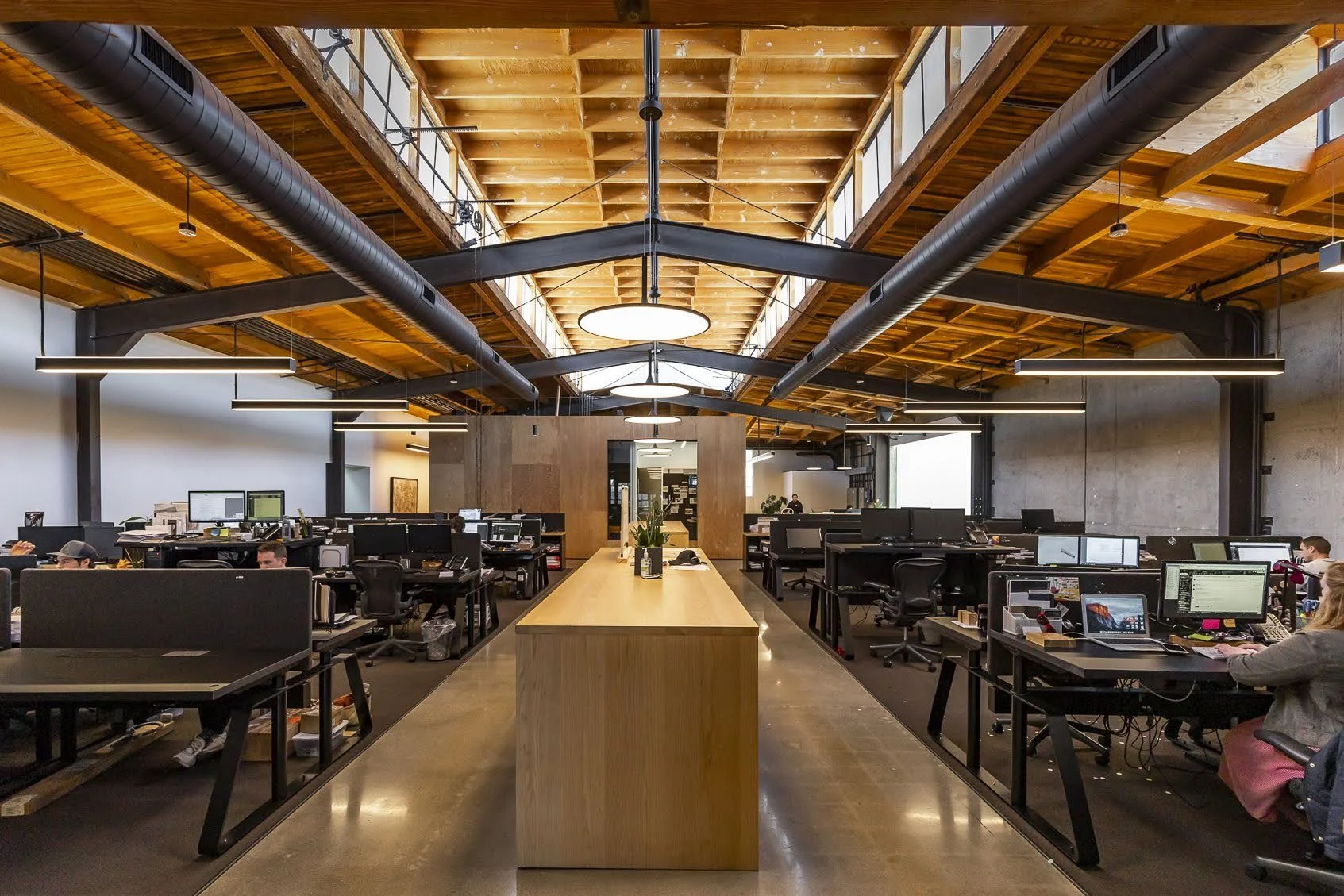 Modern open-concept office space with high wooden ceiling, exposed ductwork, and rows of desks with computers and employees.