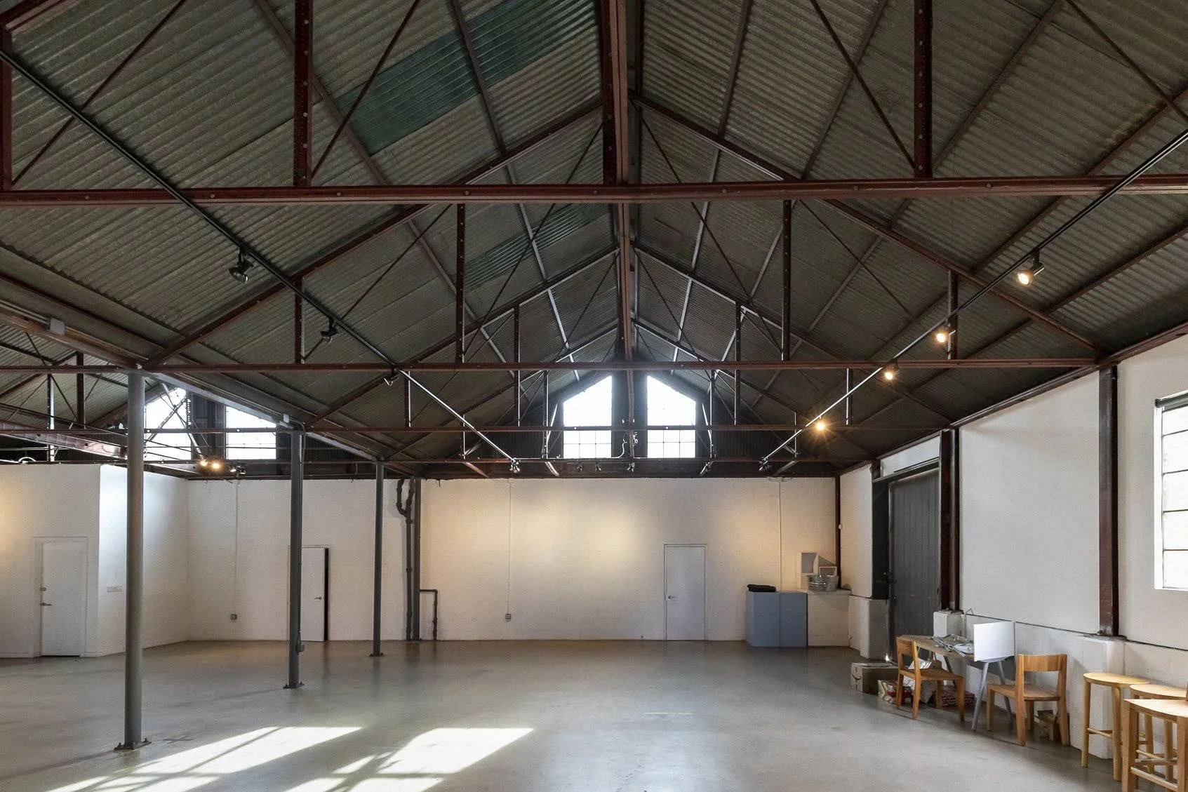 Empty industrial-style room with high vaulted ceiling, exposed metal beams, and natural light from windows, with minimal furniture and equipment on the right side.
