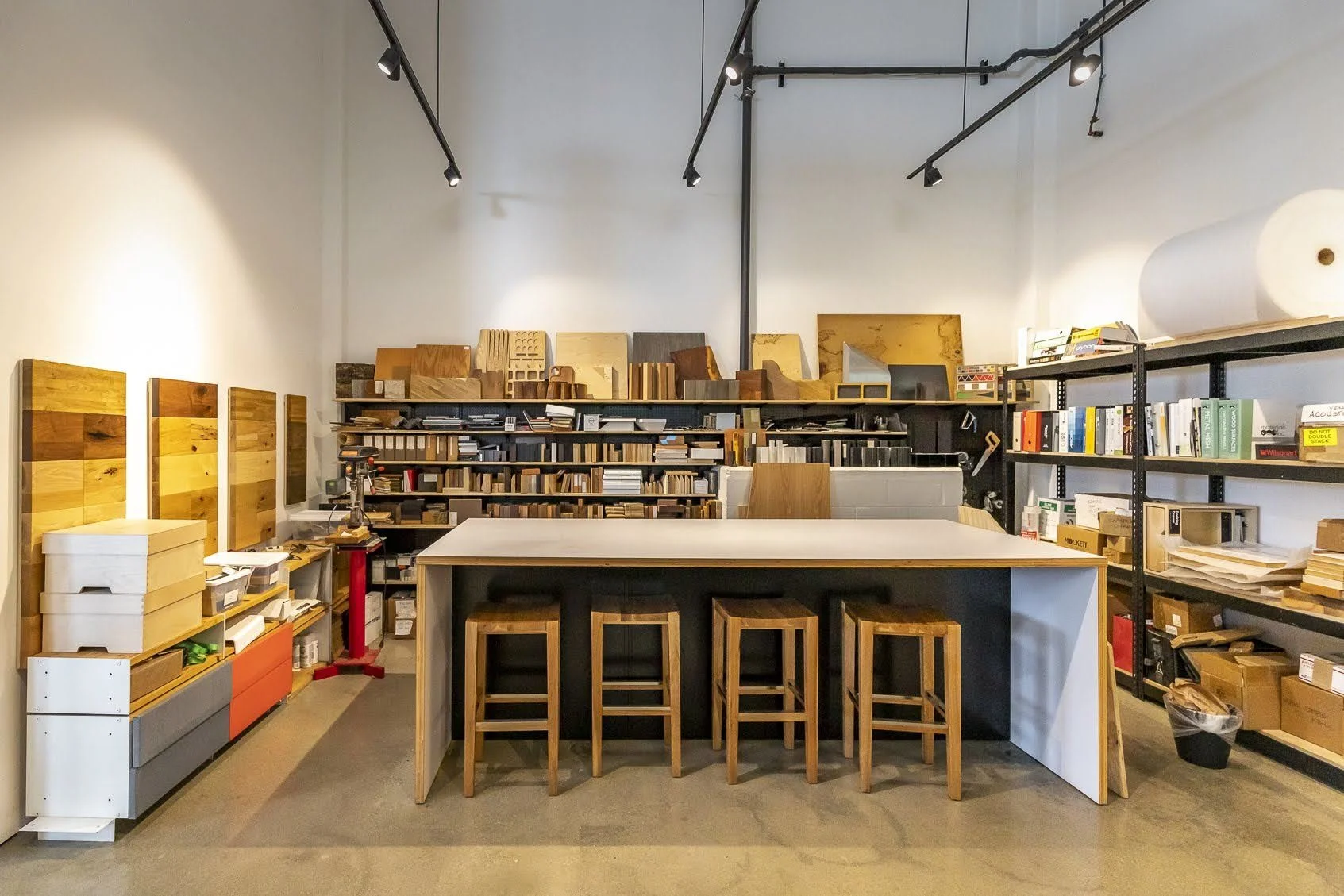 A woodworking or carpentry workshop with shelves and workbenches filled with wood samples, tools, and books. There are wooden stools around a large central table, and the walls display various wood pieces and samples.