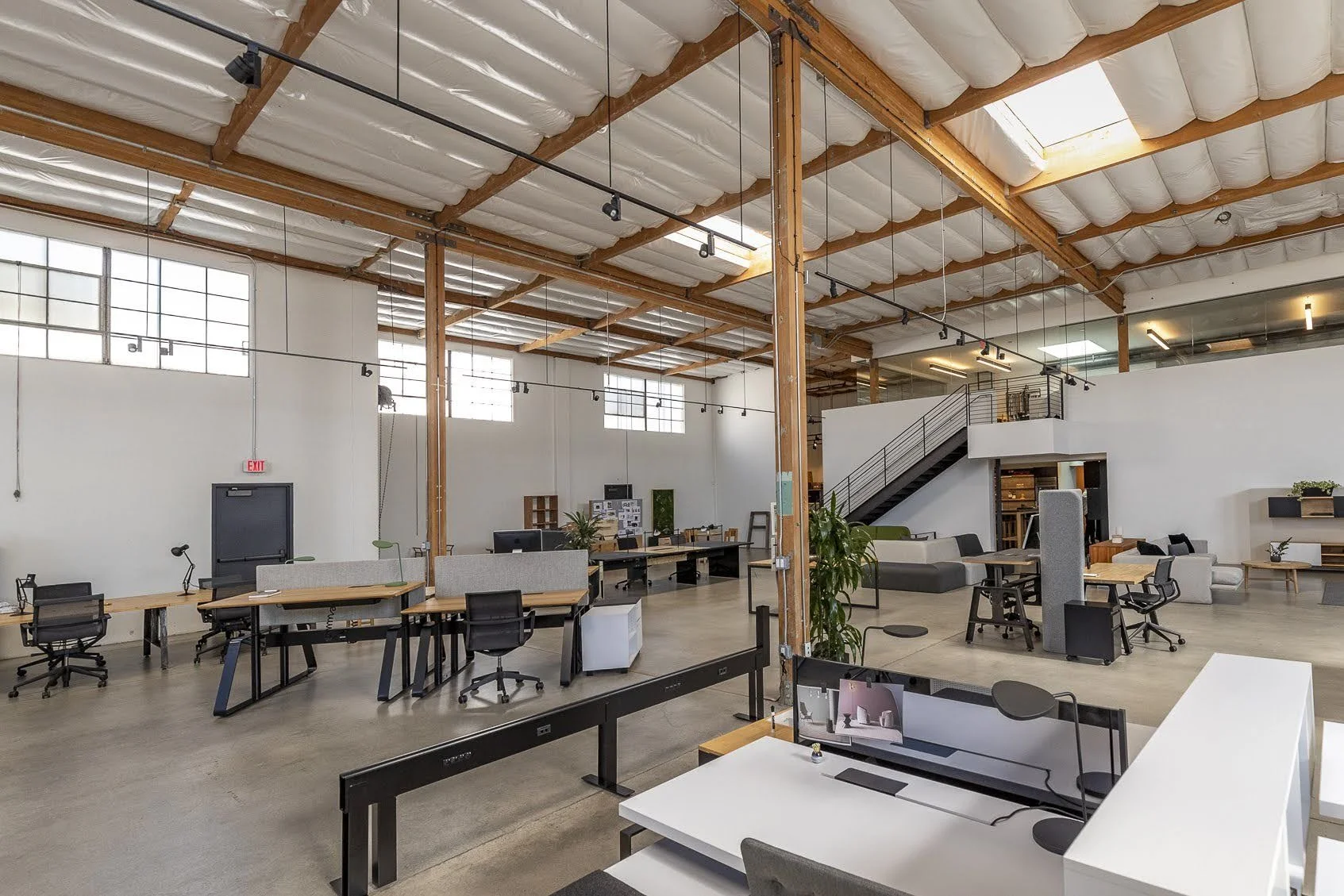 Modern open-plan office with high ceilings, large windows, and exposed wooden beams. Contains desks, chairs, computers, plants, and a staircase leading to an upper level.