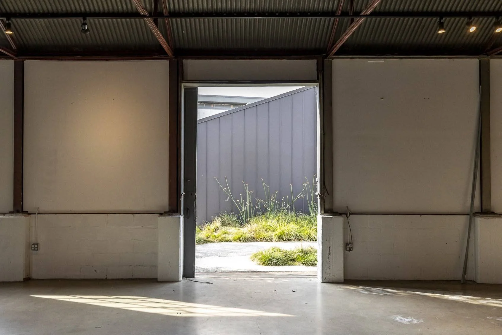 Interior of an unfinished building with an open door leading outside, showing a garden with grass and plants.