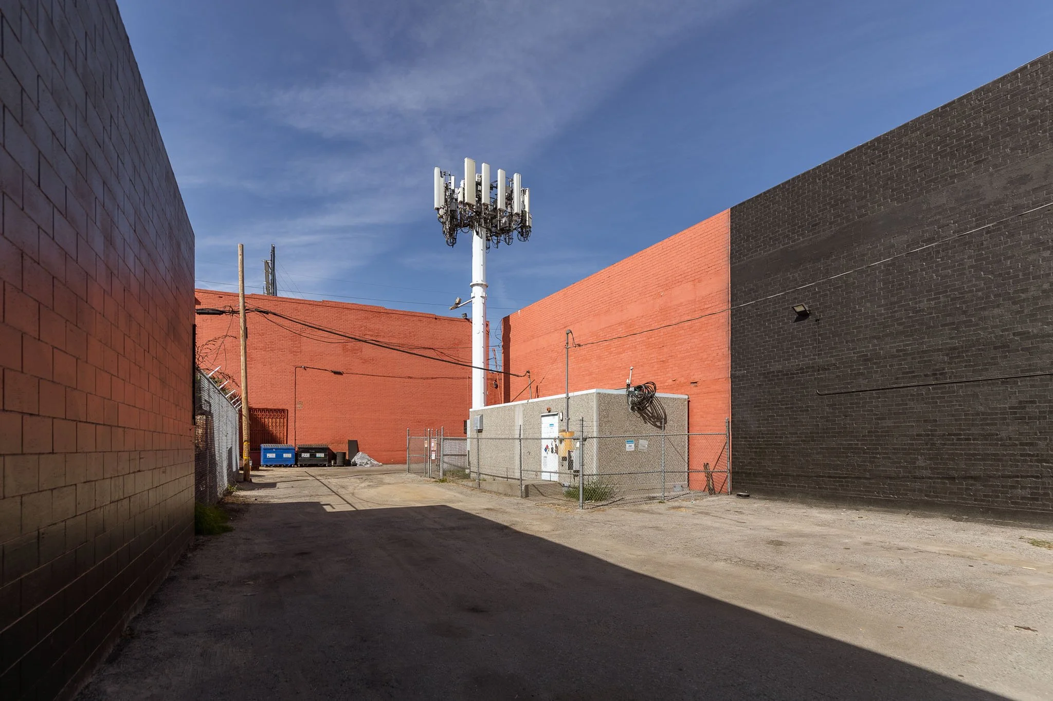 Alleyway with red and black brick walls, utility pole, small building with fence, and cell tower against a blue sky.