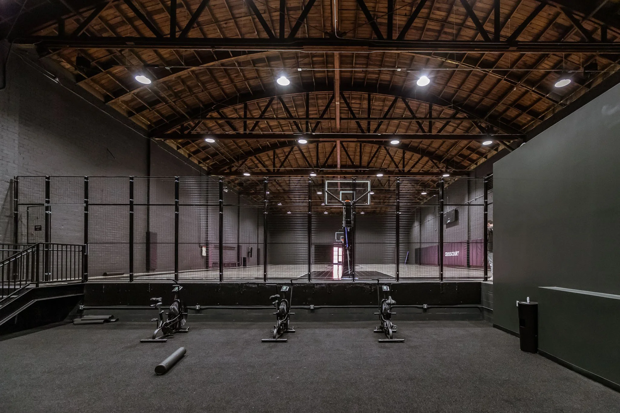 An indoor basketball court with a hoop, enclosed by a black fence, and exercise bikes on the carpeted floor in the foreground.