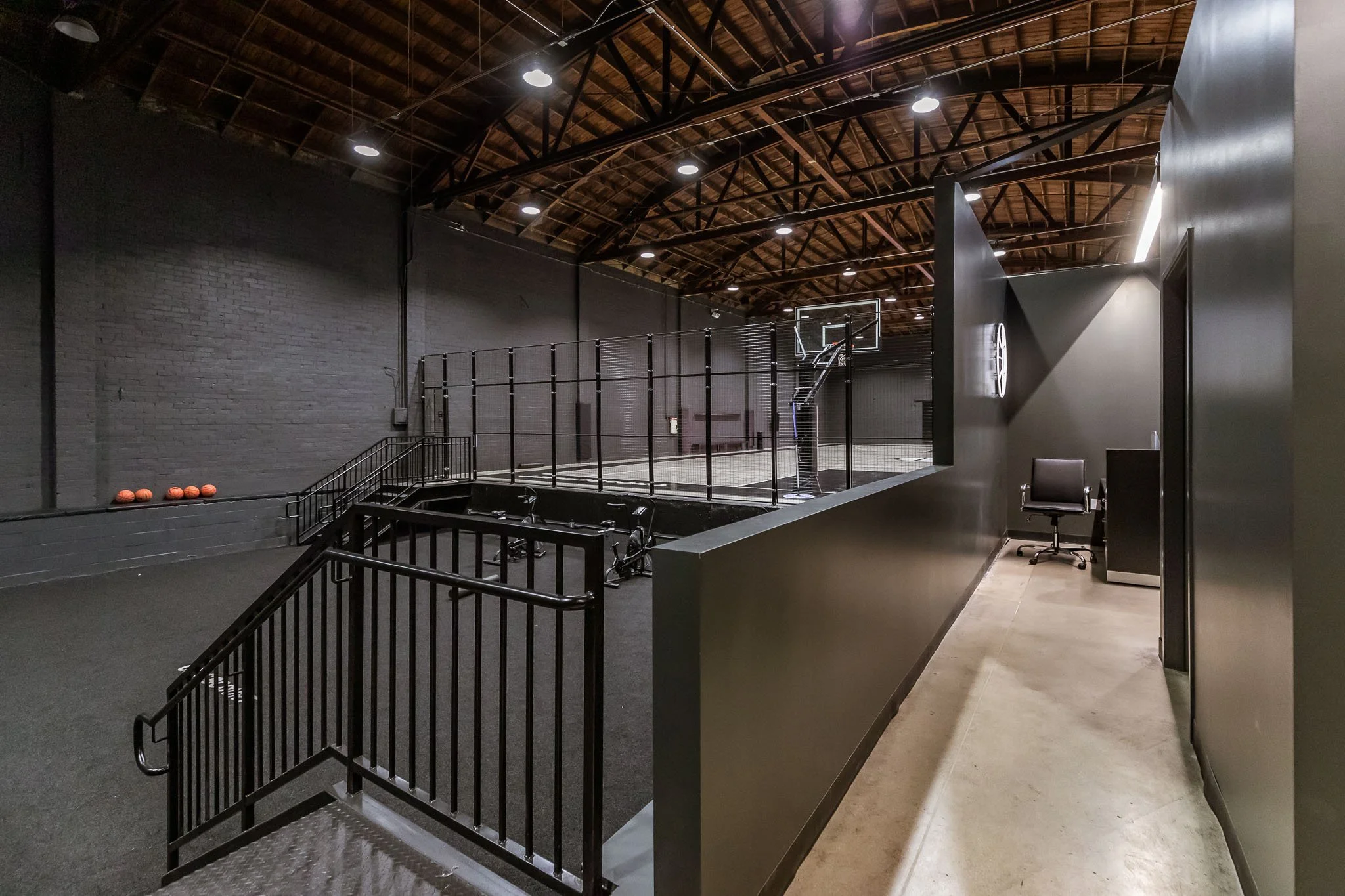 Indoor basketball court with black walls, a basketball hoop, and a row of orange basketballs on the floor in the background. There is a small balcony with a black railing overlooking the court and a side room with a chair and desk.