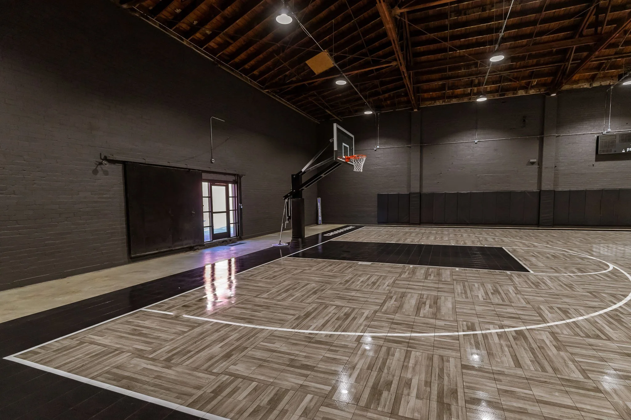 An indoor basketball court with a wooden floor, black rebound wall padding, basketball hoop, and high ceiling with exposed beams and lighting.