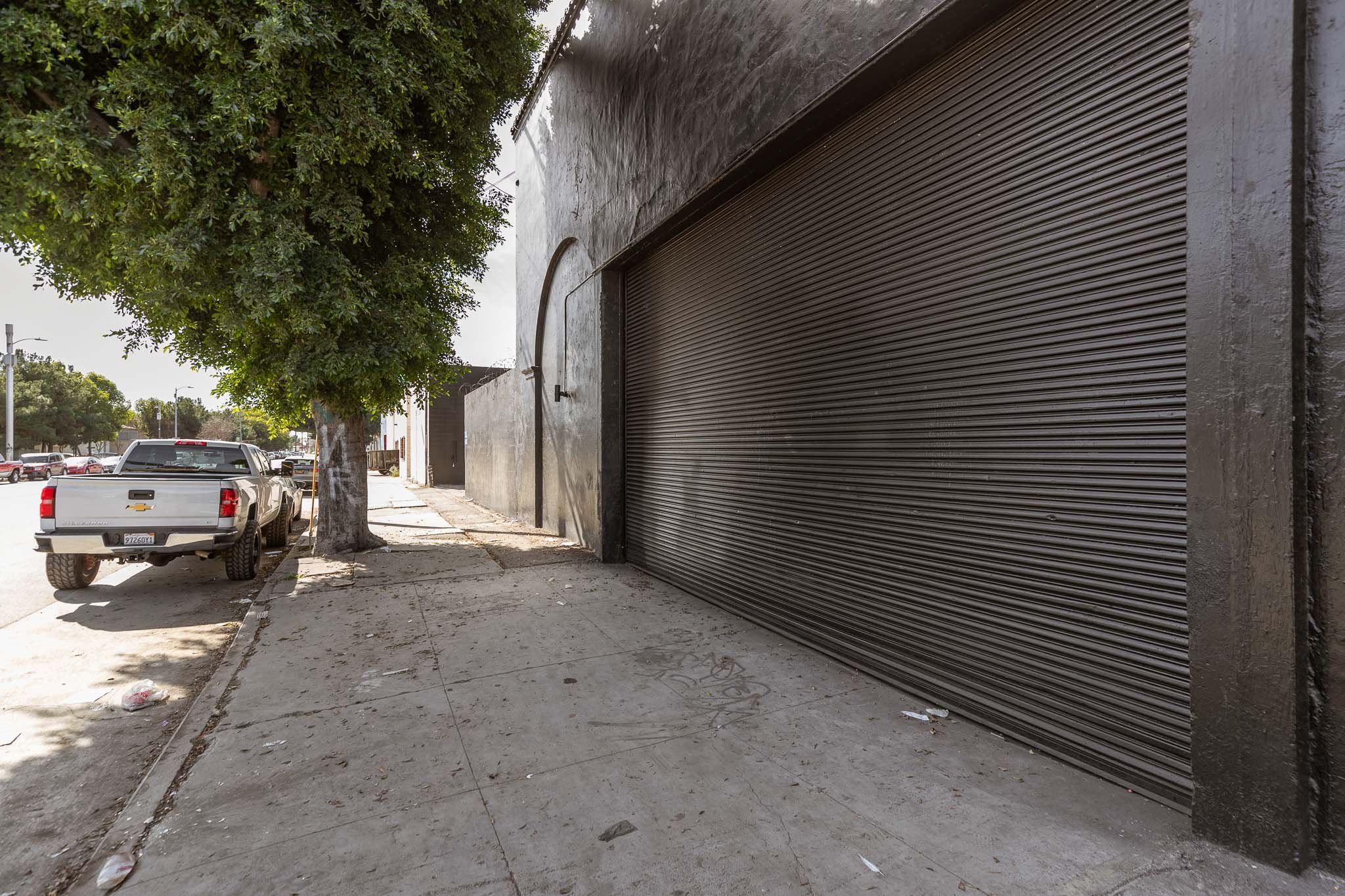 Empty sidewalk with a closed metal roll-up gate, a parked pickup truck, a large tree, and buildings on a sunny day.