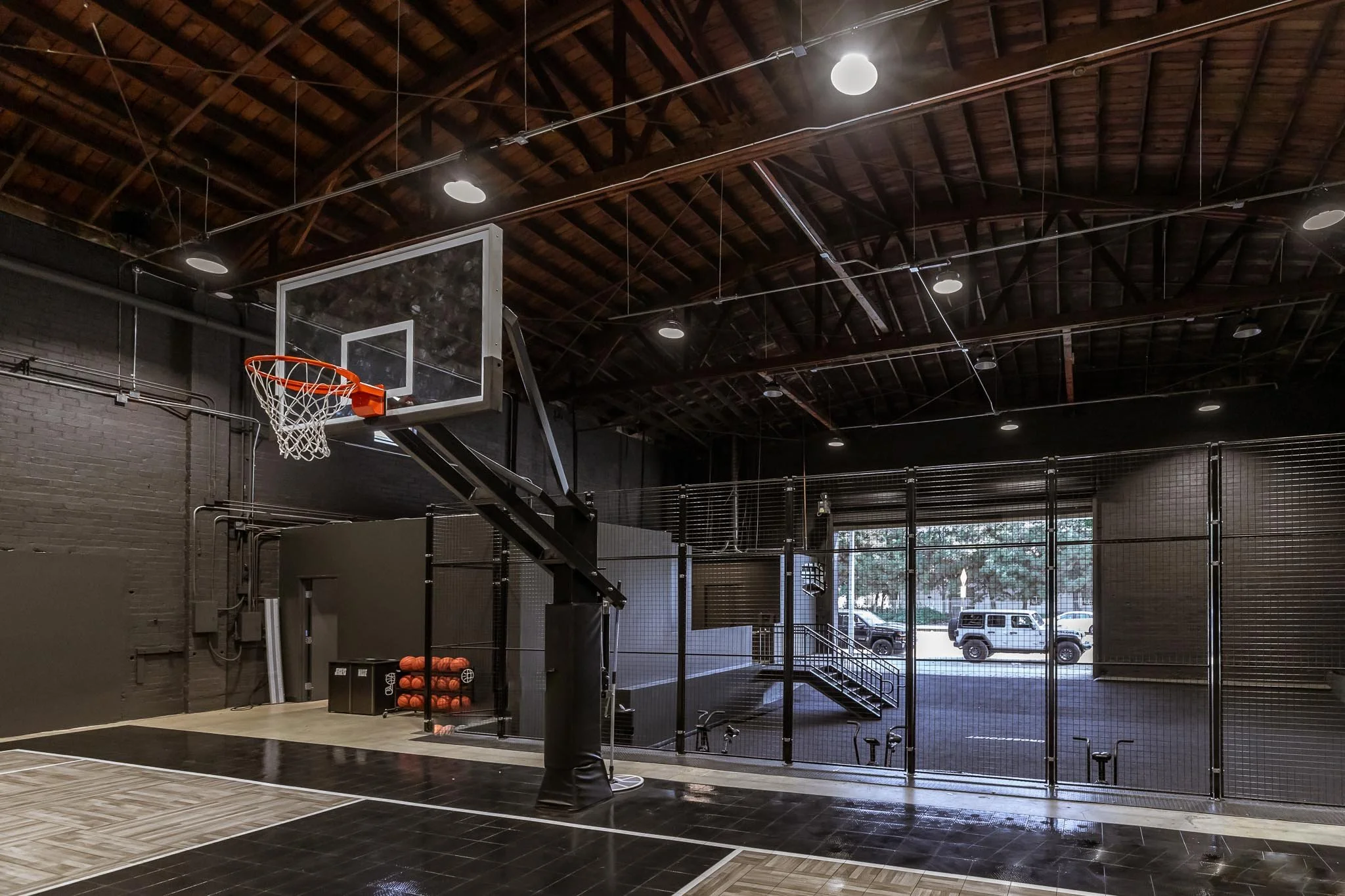 Indoor basketball court with a basketball hoop, a basket of orange balls, and a large open space with a chain-link fence and a view of parked vehicles outside.