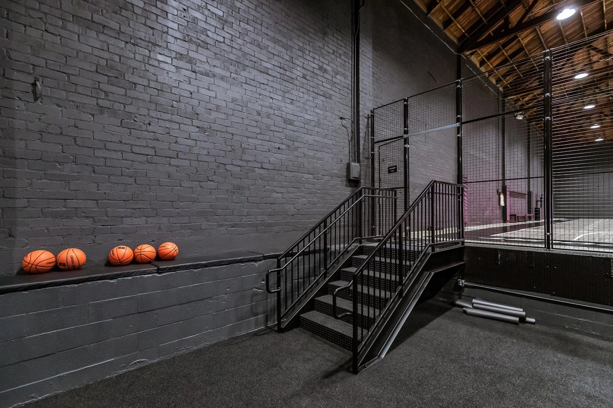 Indoor basketball court with orange basketballs on a ledge, black stairs with handrails, and a fenced court area in the background.
