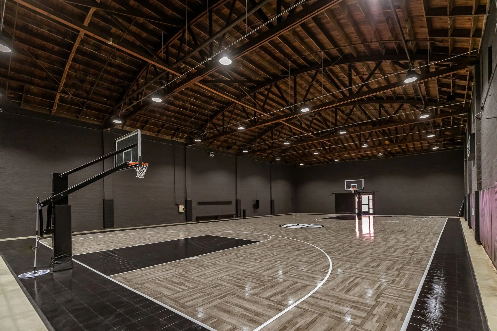 Indoor basketball court with wooden flooring, black wall, and high wooden ceiling with suspension lighting. Two basketball hoops are visible at opposite ends of the court.