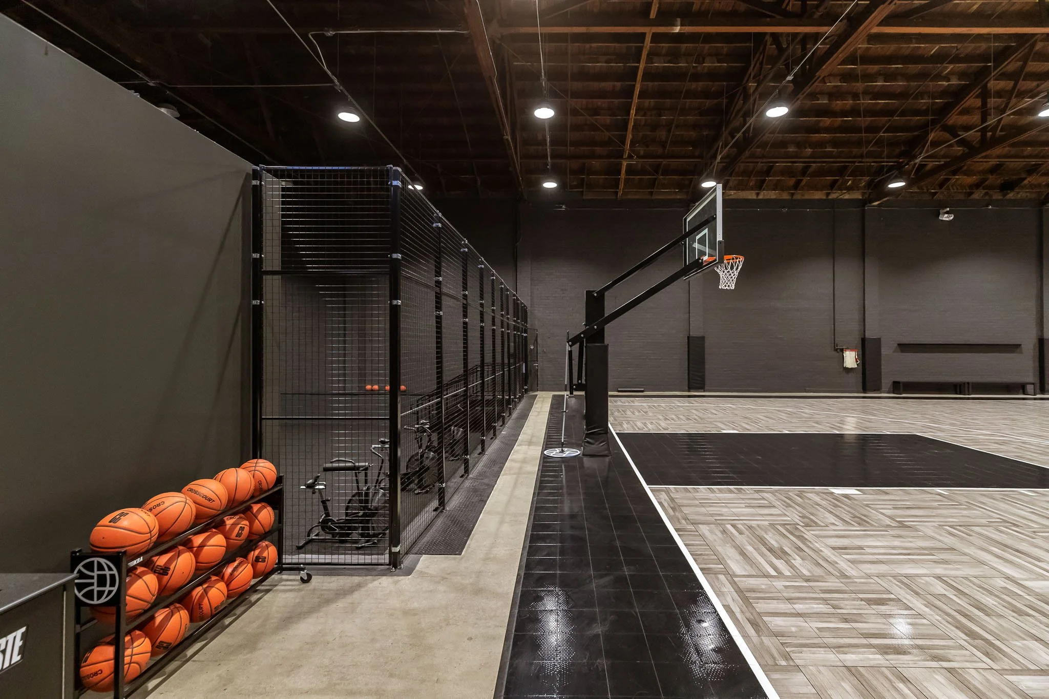 Empty indoor basketball court with black walls, wooden floor, basketball hoops, and basketballs stored on shelves.
