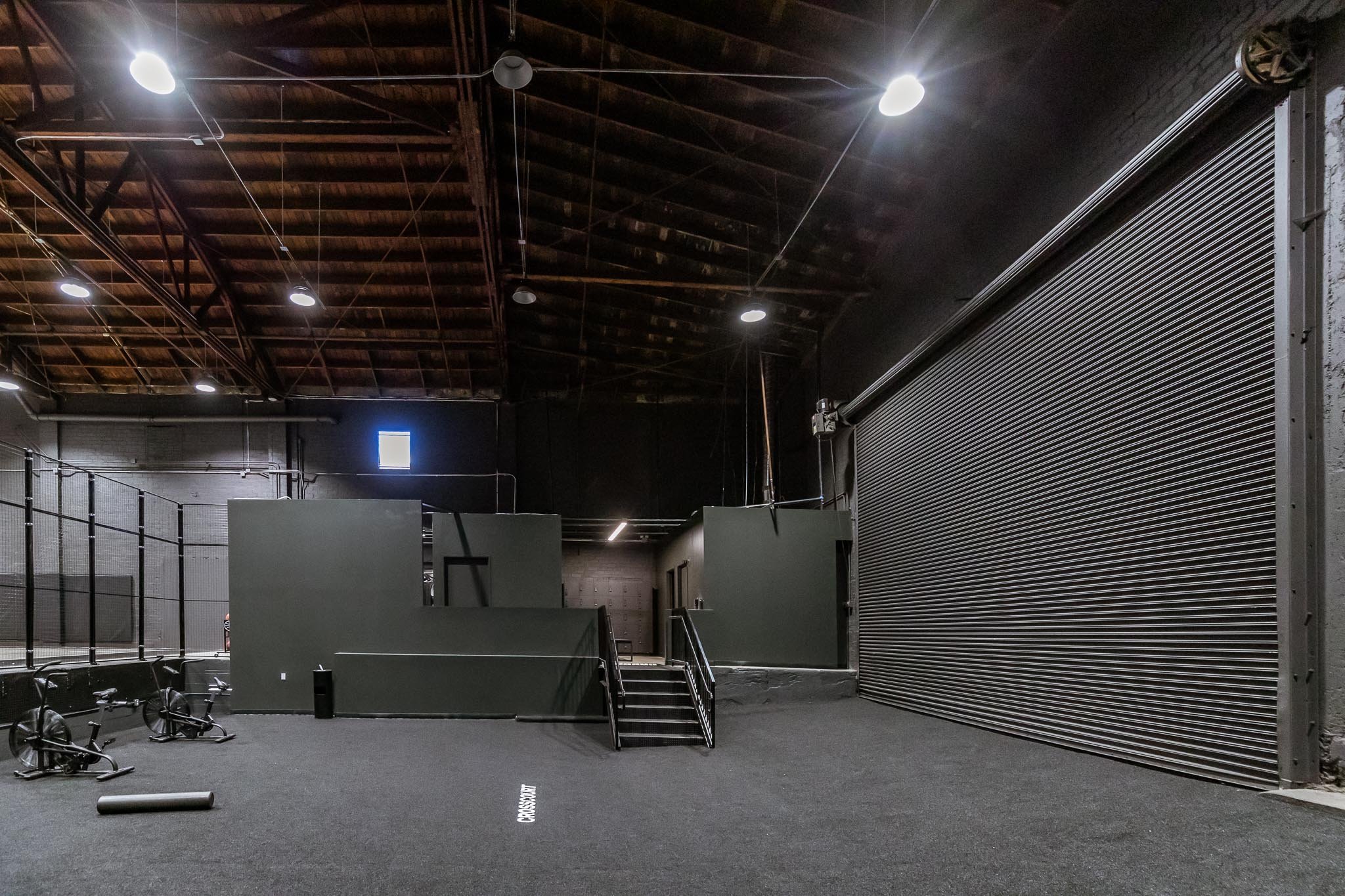 Empty indoor gym with black rubber flooring, stationary bikes, green walls, a large metal roll-up door, and a high wooden ceiling with hanging lights.