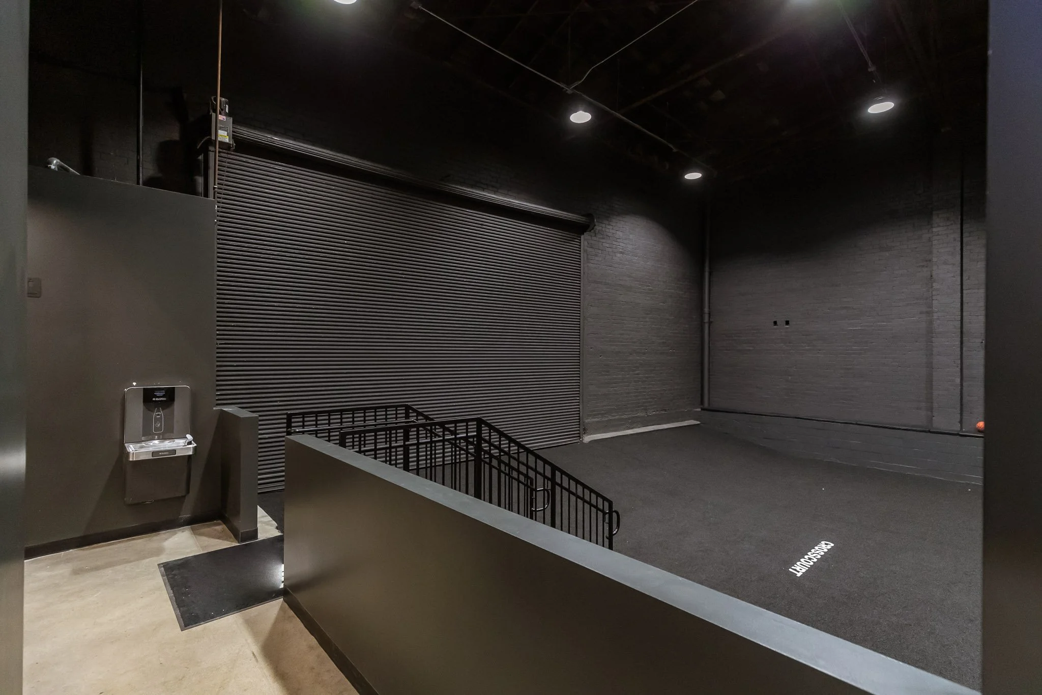 Empty indoor basketball training court with black flooring, black brick walls, and a closed metal roller shutter door.