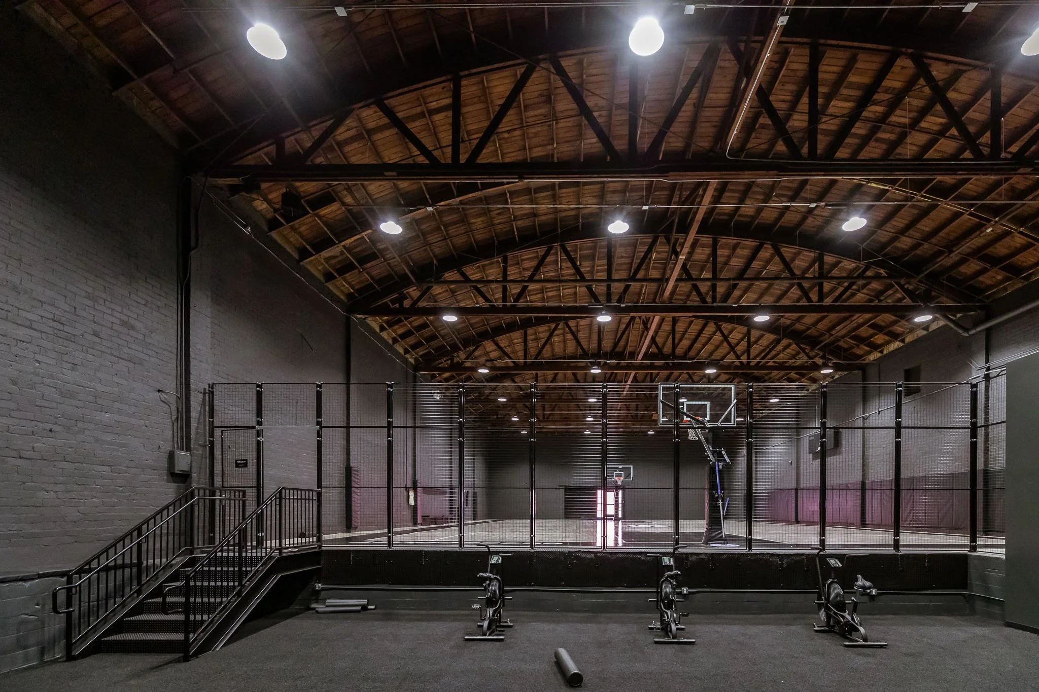Indoor basketball court with two hoops, surrounded by black fencing, illuminated by ceiling lights, with exercise bikes at the foreground, and a black brick wall with visible piping and a staircase on the left.