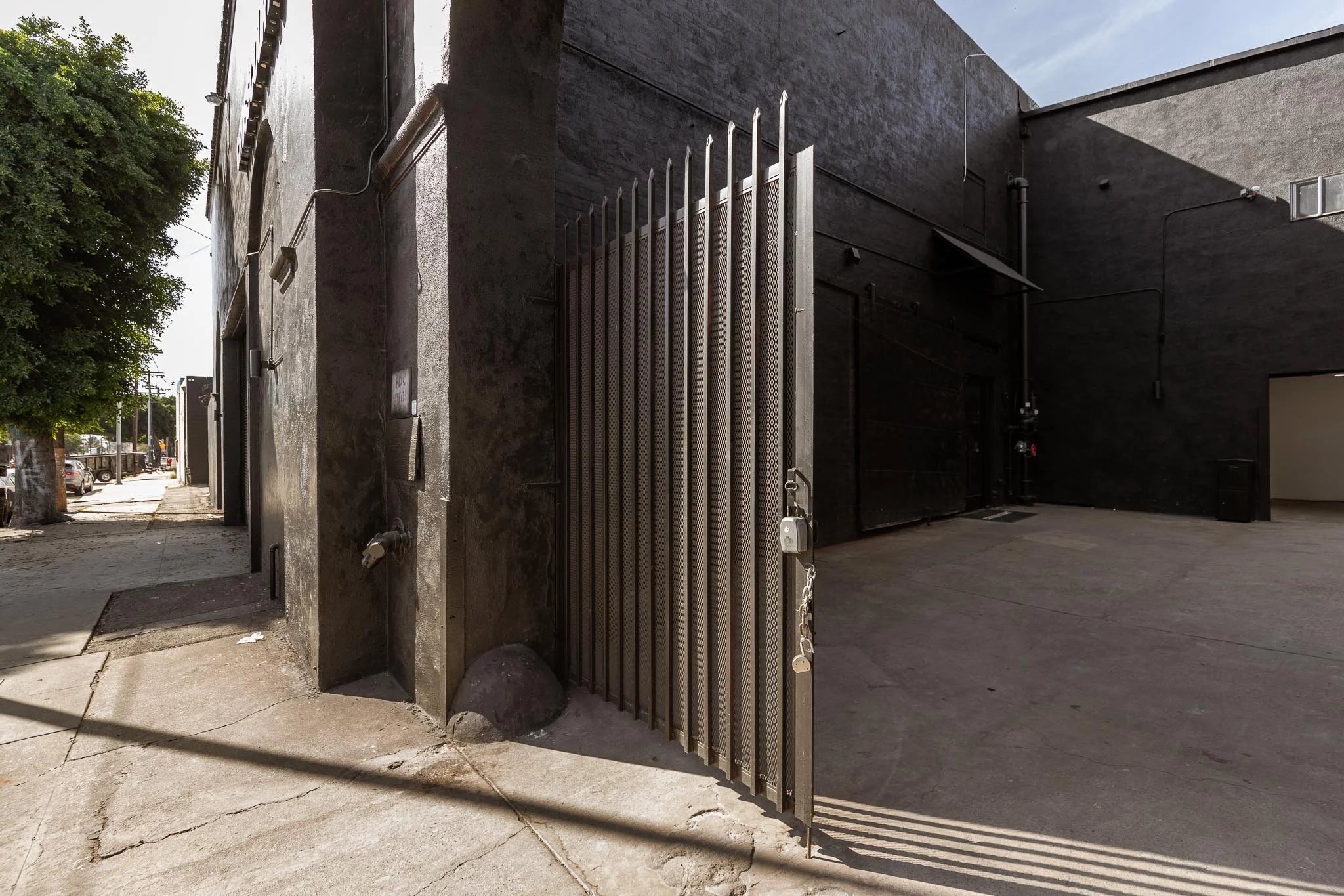 Black building with metal gate and padlock on an urban sidewalk, with trees and parked cars in the background.