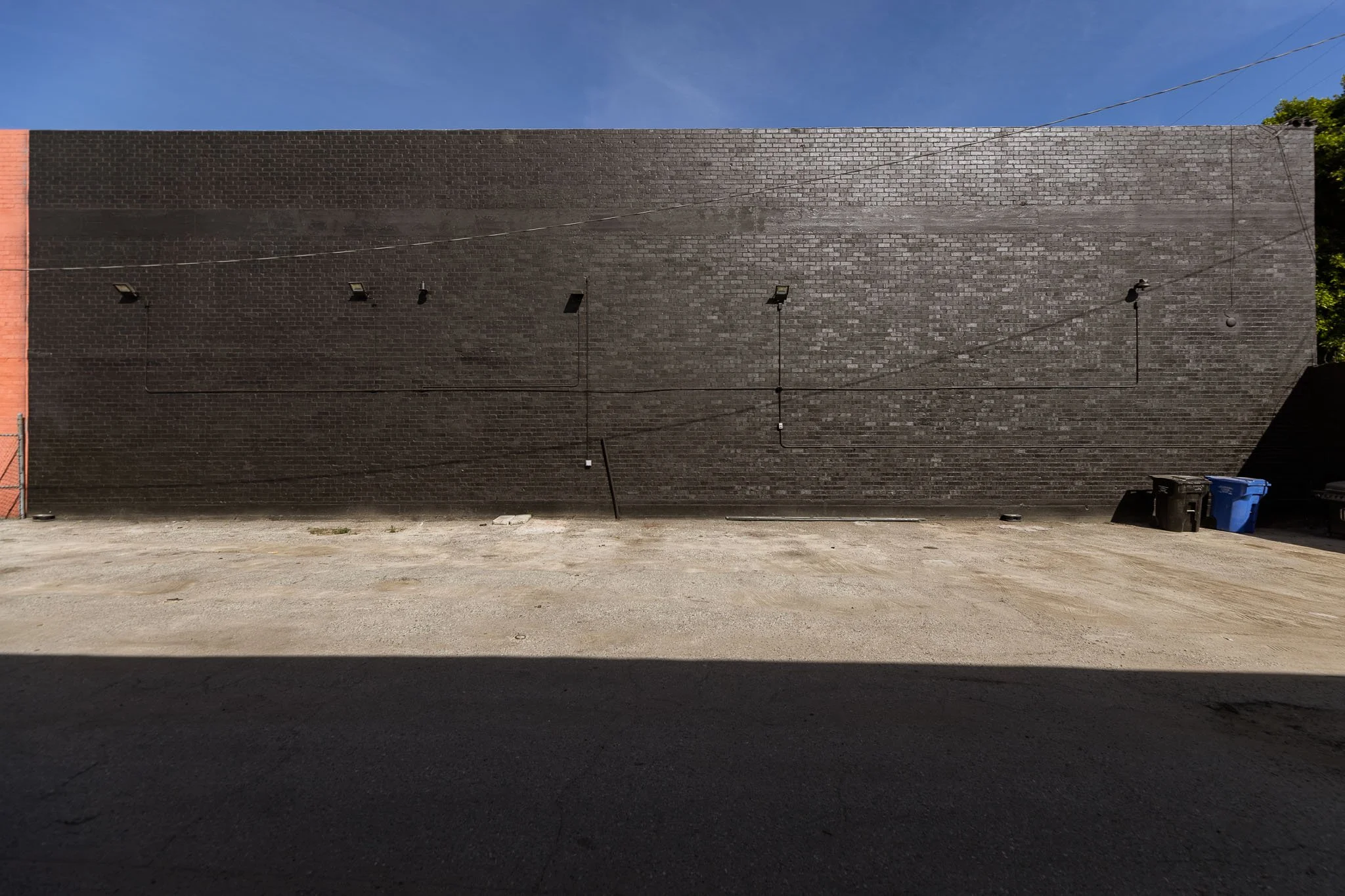 Blank brick wall with four mounted lights and electrical wiring, with a dirt ground and two trash bins in front.