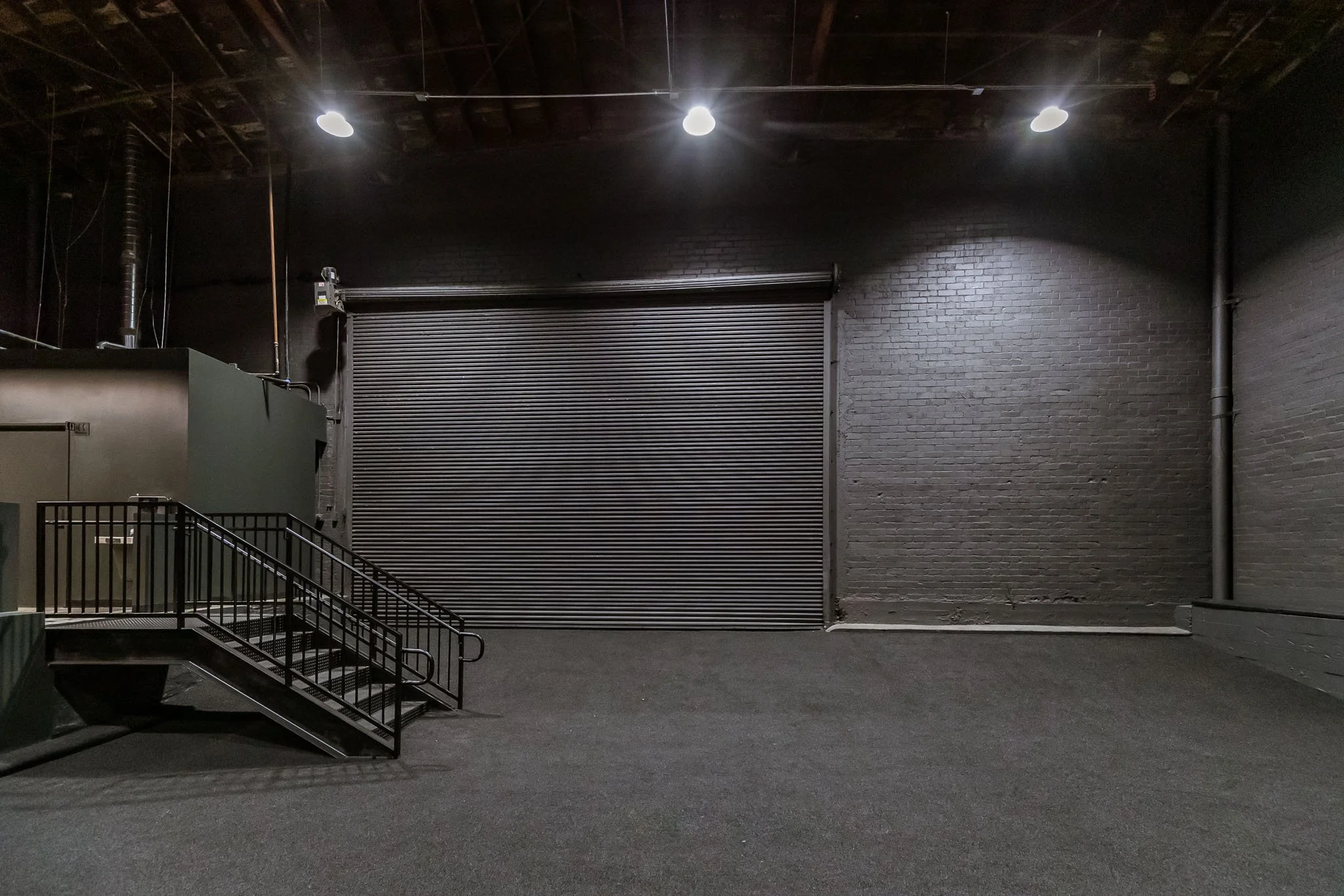 Empty black room with a metal roll-up door, a small staircase with black railings, gray flooring, black brick walls, and overhead lighting