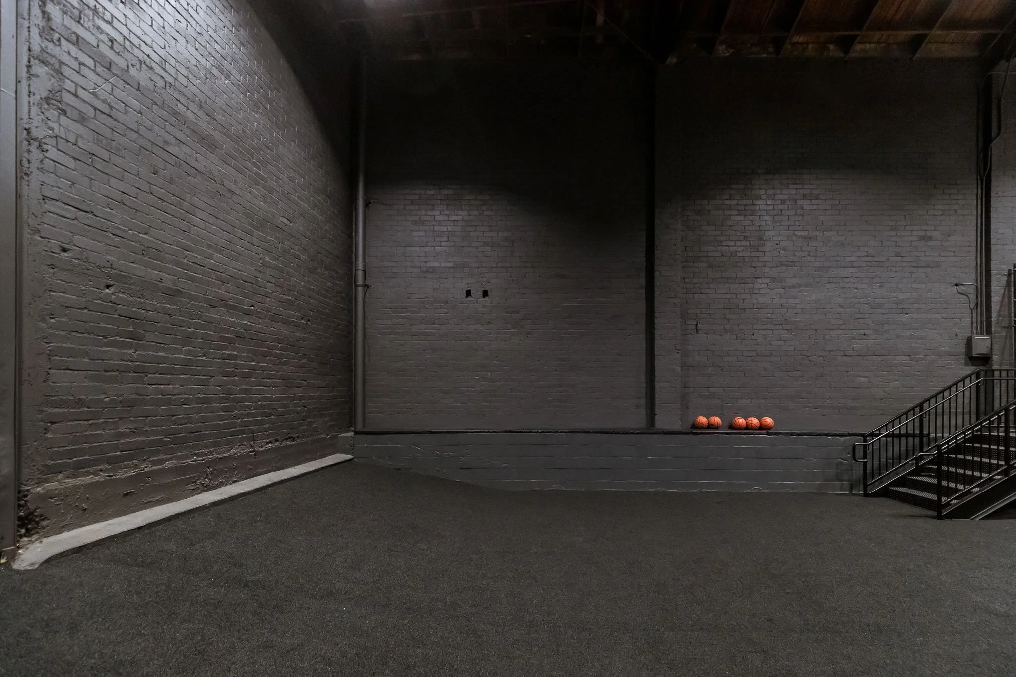 An empty interior space with dark brick walls, a carpeted floor, and a small set of metal stairs on the right side. Four basketballs are resting on a raised platform against the wall.