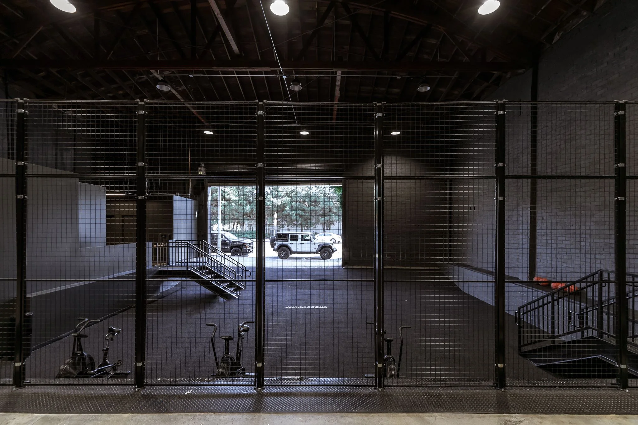 Empty indoor gym with black fencing, exercise bikes in foreground, stairs, and open garage door showing parked cars outside.
