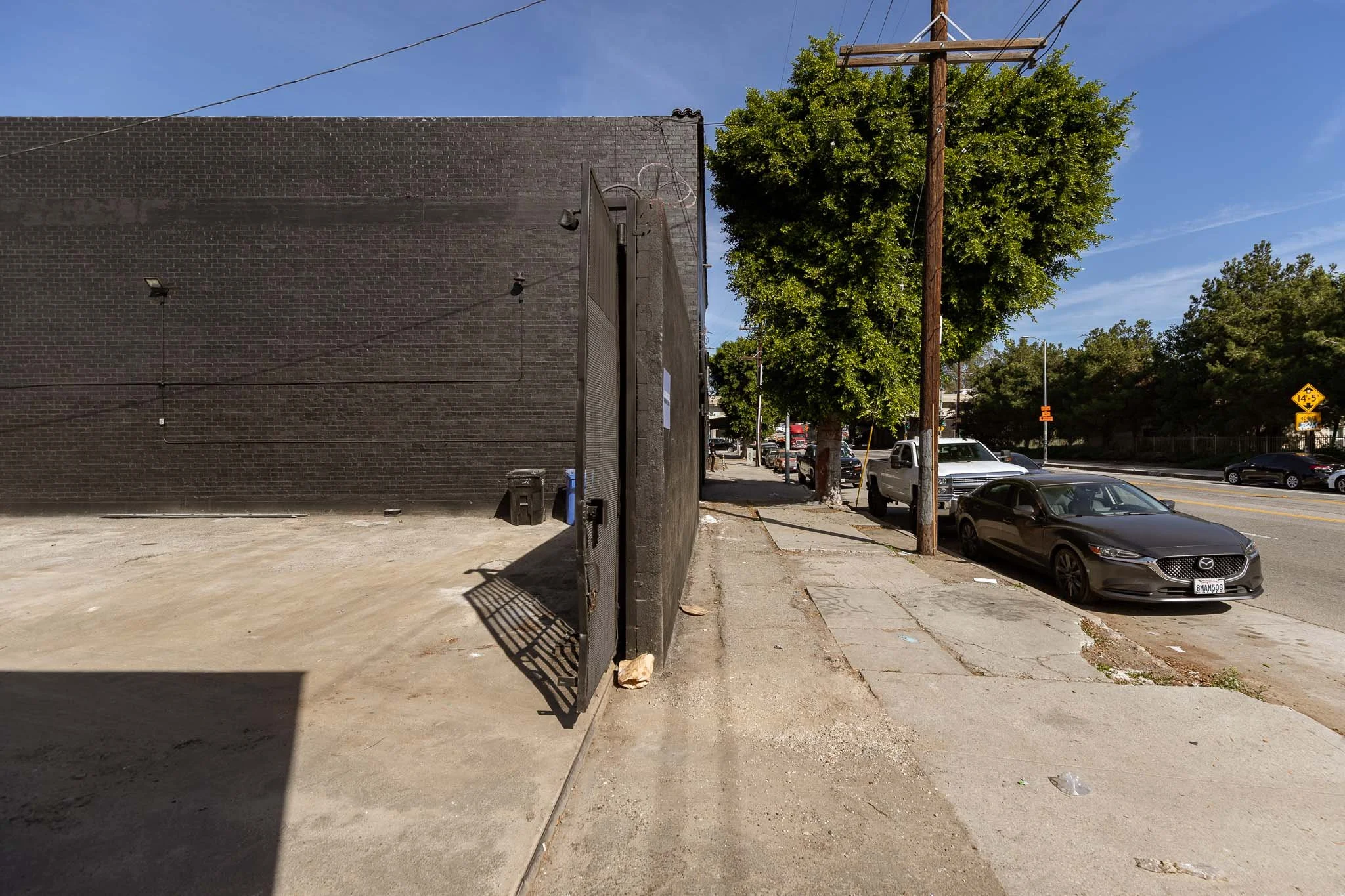 Urban street scene showing a sidewalk adjacent to a black brick building with a black gate, parked cars including a gray Mazda, trees, utility poles, and a yellow traffic sign in the distance, under a blue sky.