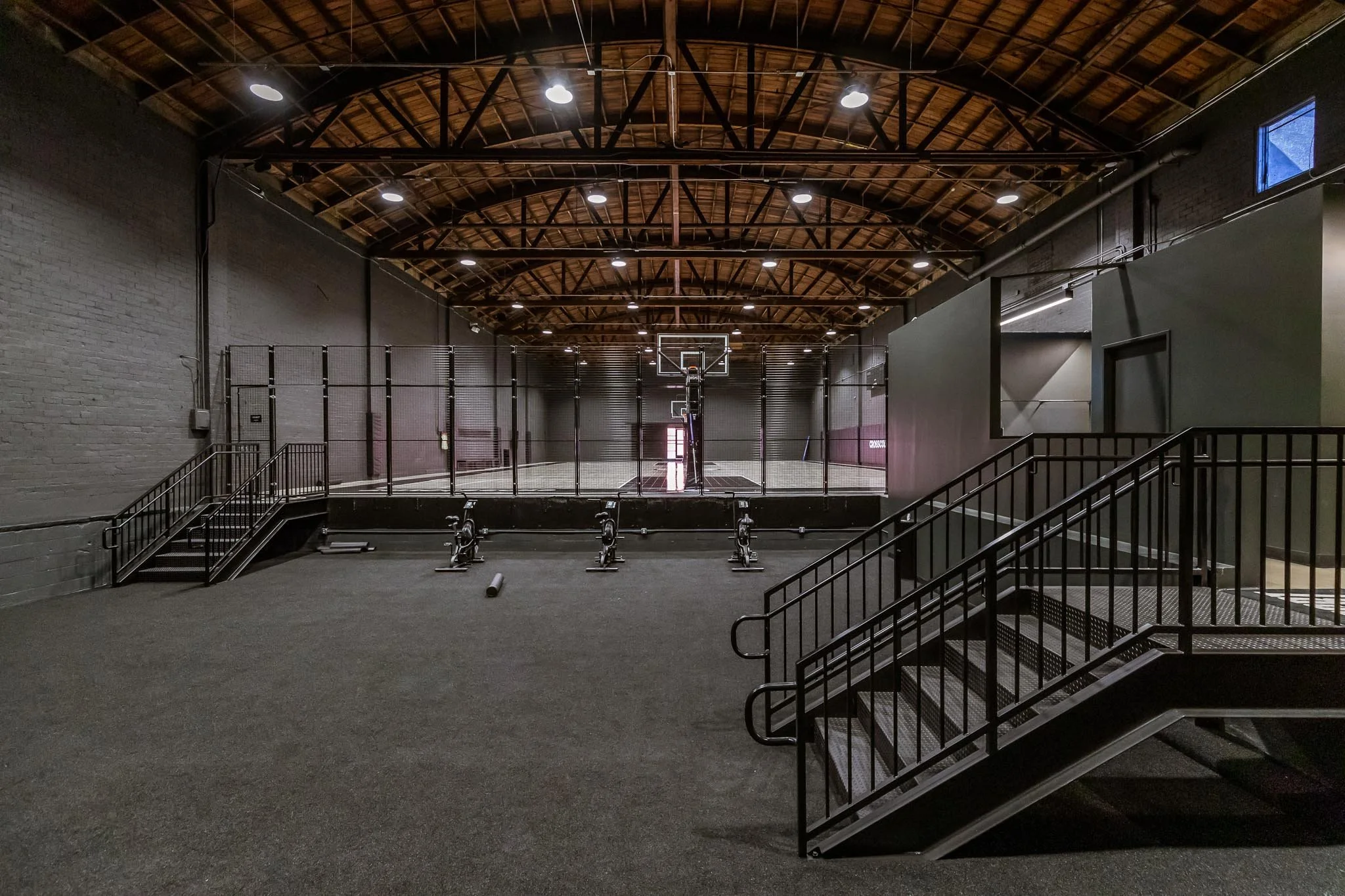 Empty indoor basketball gym with a regulation hoop, black padded floor, and a fenced volleyball court in the background, with stairs and high wooden ceiling.