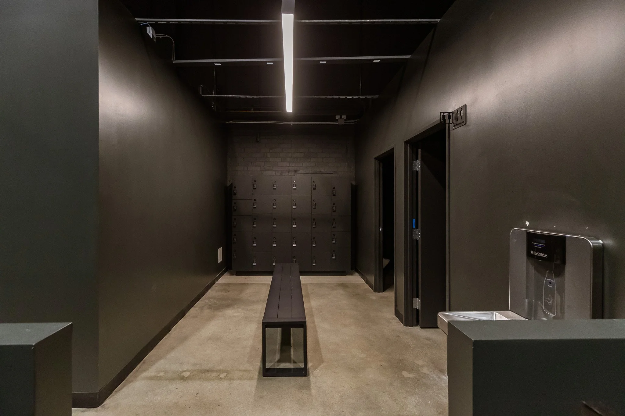 A modern room with black walls, gray lockers at the back, a black bench in the center, and a water fountain on the right side.
